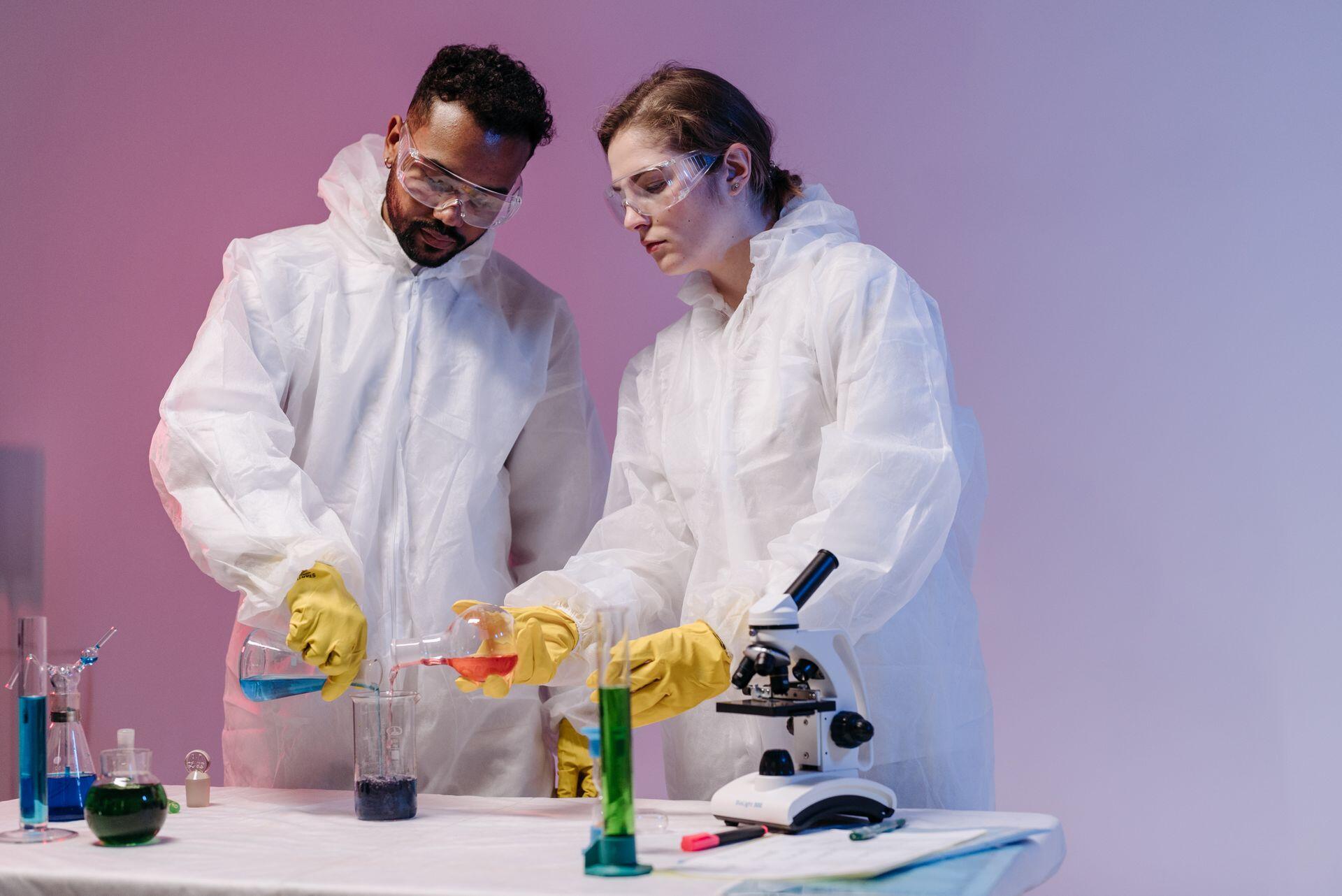 Dos personas con traje de laboratorio blanco sosteniendo frascos con líquidos de colores.