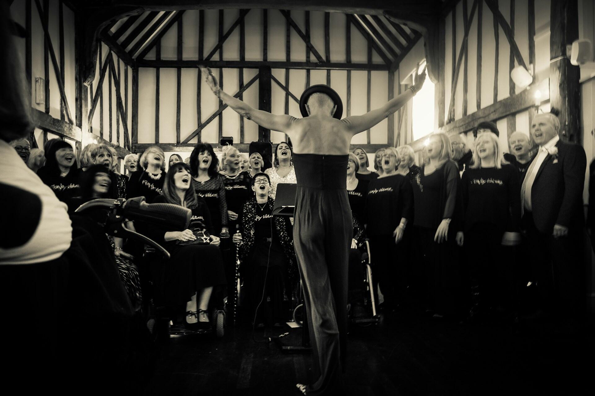 Interior de un salón de madera rústico con un coro ensayando técnicas de vocalización grupal, por Colin Michael.