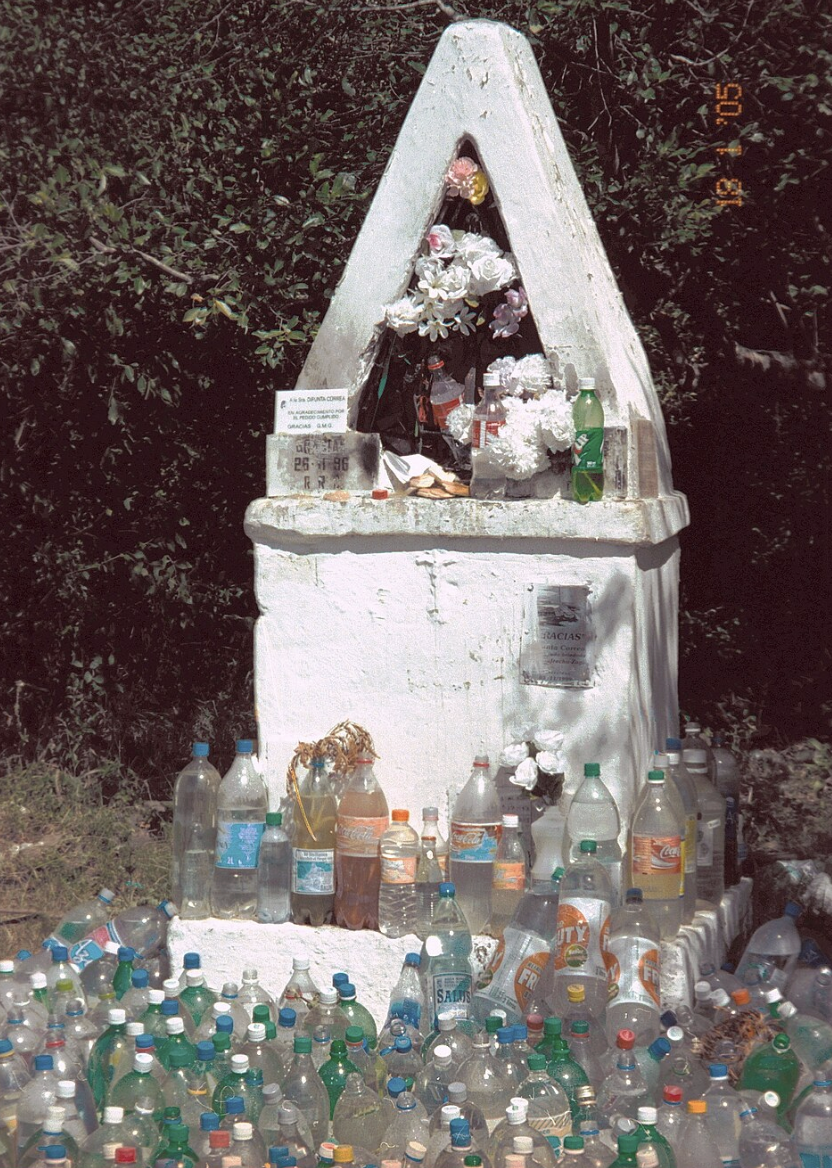 Pequeño altar blanco en forma de pirámide con flores y botellas de agua en la base, dedicado a la Difunta Correa. Fuente: Wiki Commons.