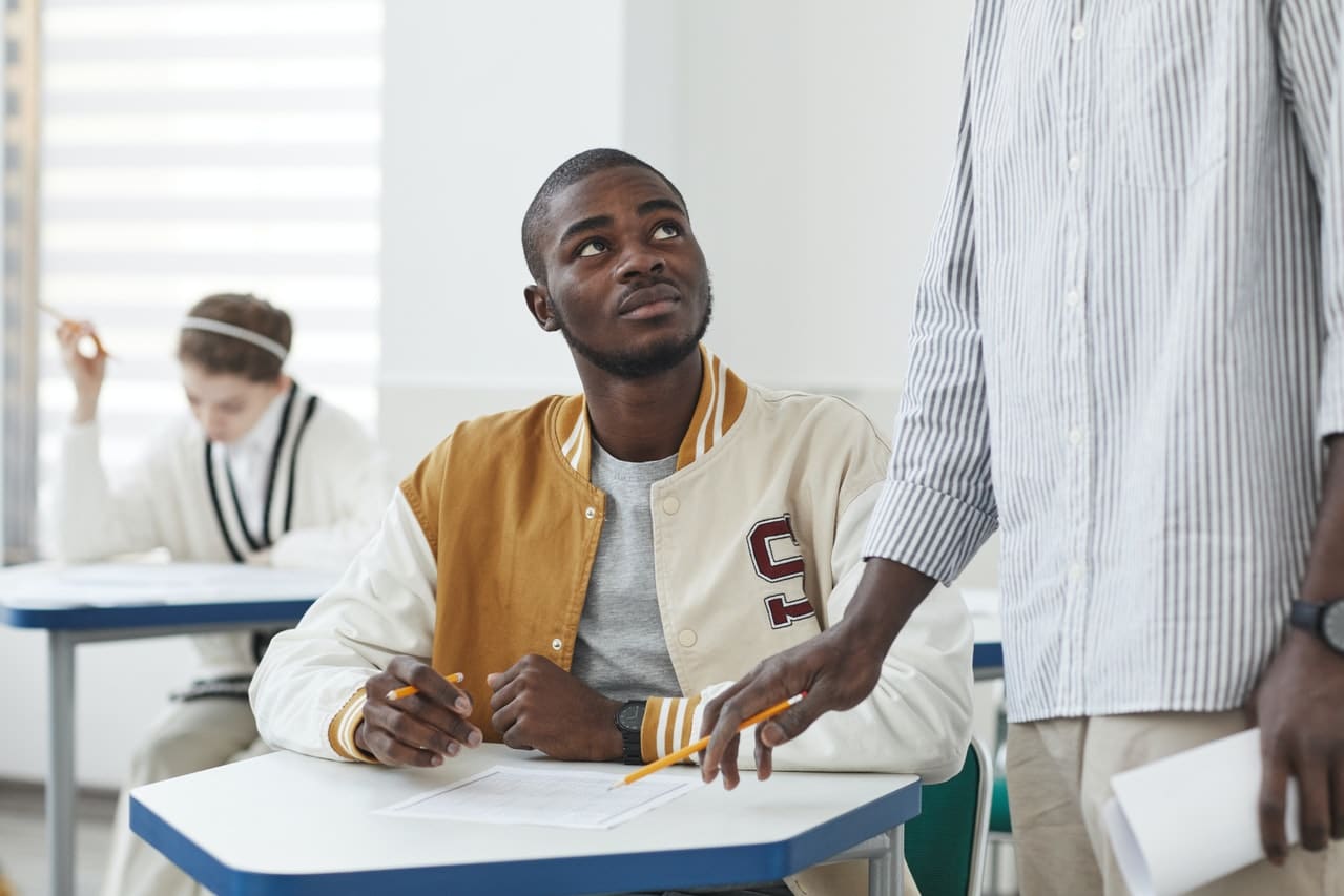 Schüler sitzt an Tisch und richtet Blick auf Lehrer