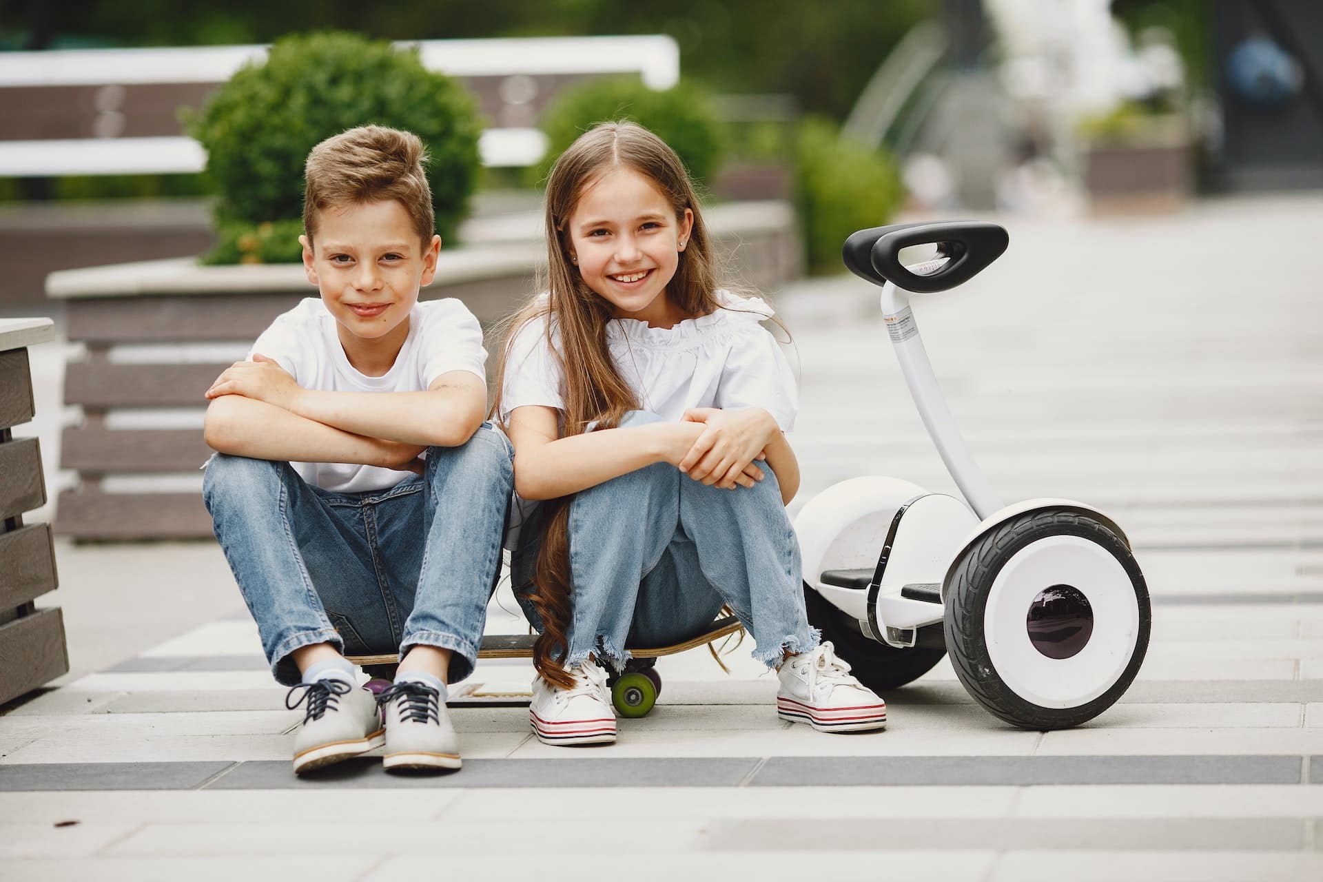 Zwei Volksschulkinder sitzen auf einem Skateboard auf der Straße und grinsen in die Kamera.