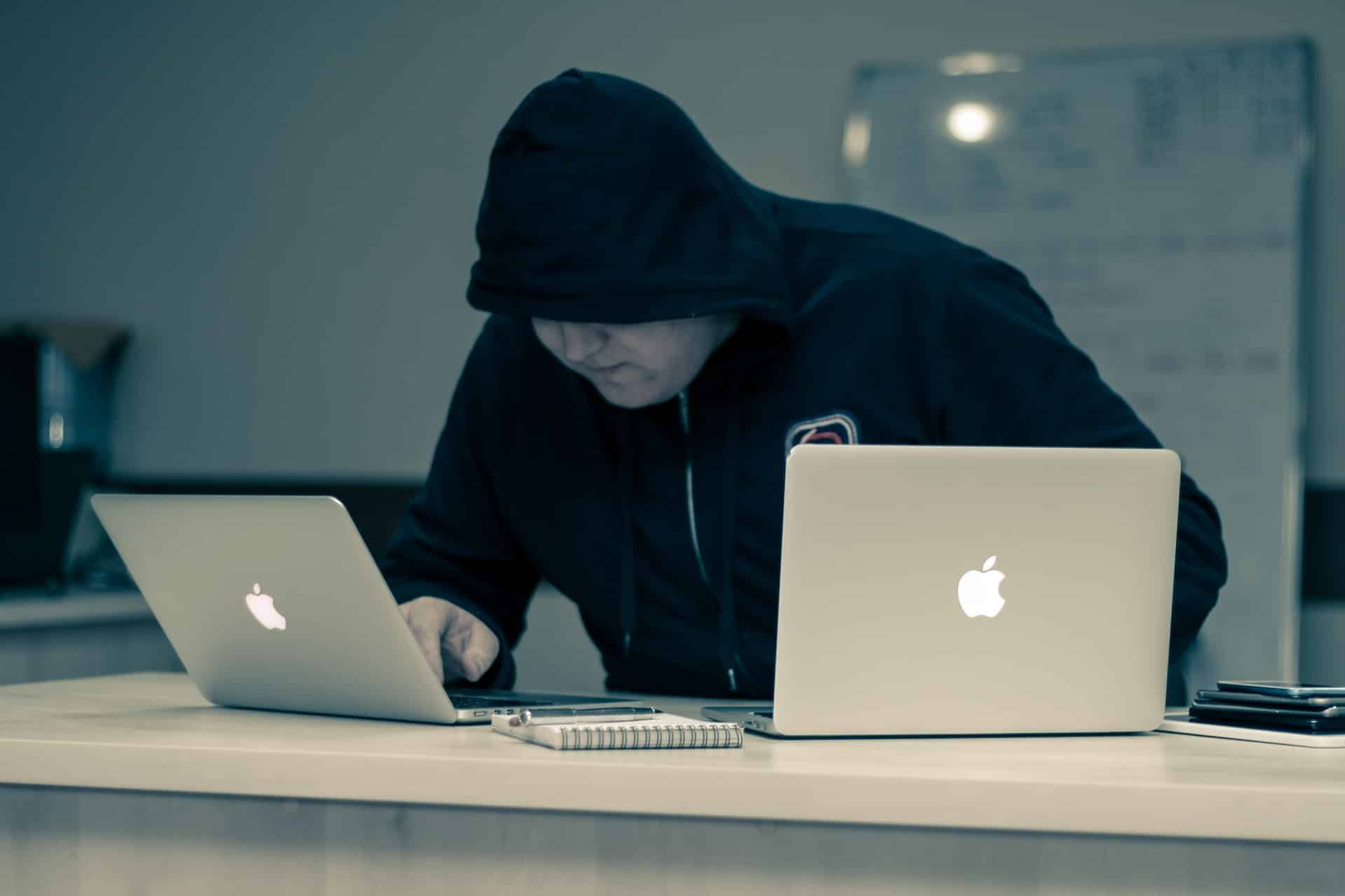 Ein Mann mit einem schwarzen Pullover bekleidet sitzt auf einem Tisch mit zwei Laptops vor sich.