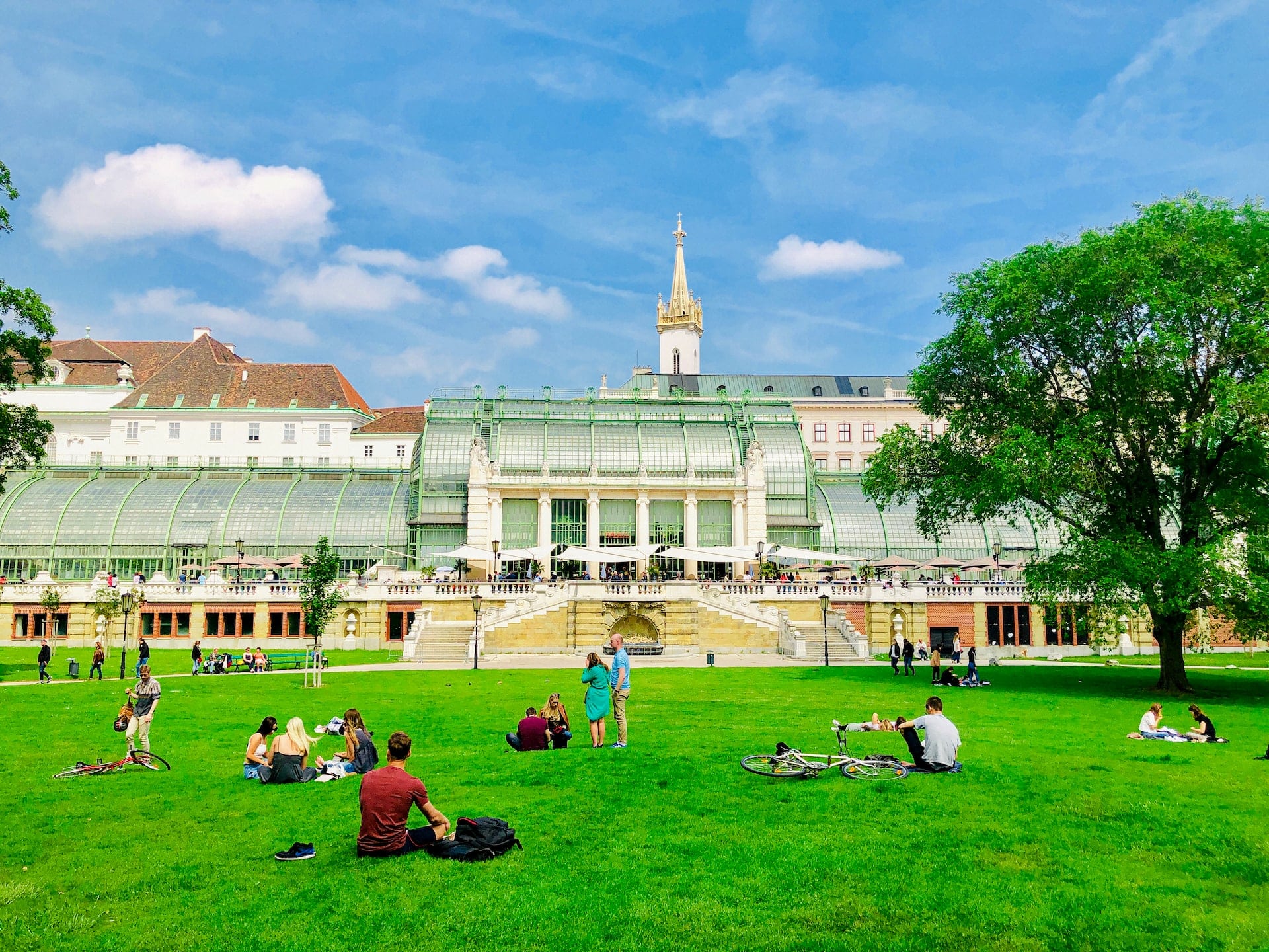 Der Burggarten in Wien mit dem Palmenhaus im Hintergrund und Menschen auf der Wiese sitzend in sehr grellen Farbtönen.