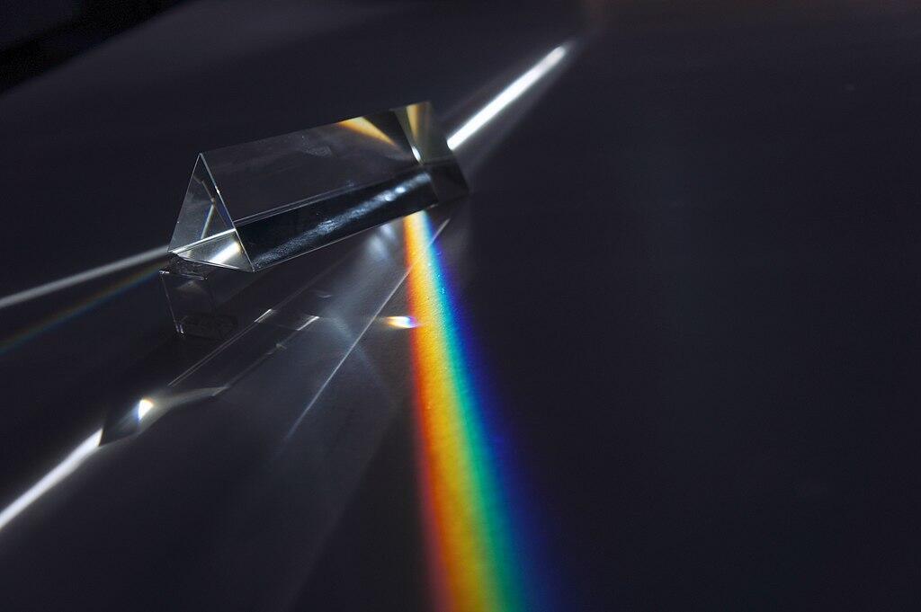 Ein in Regenbogenfarben aufgeteilter Lichtstrahl geht durch ein Prisma und wird wieder zu weißem Licht gebündet.