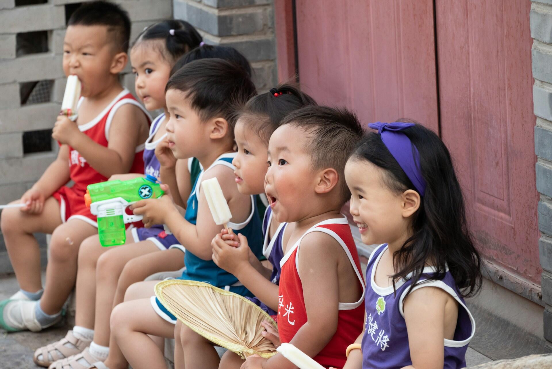 Young children wearing summer clothing sit in a row, in front of a red door, eating snacks and smiling.