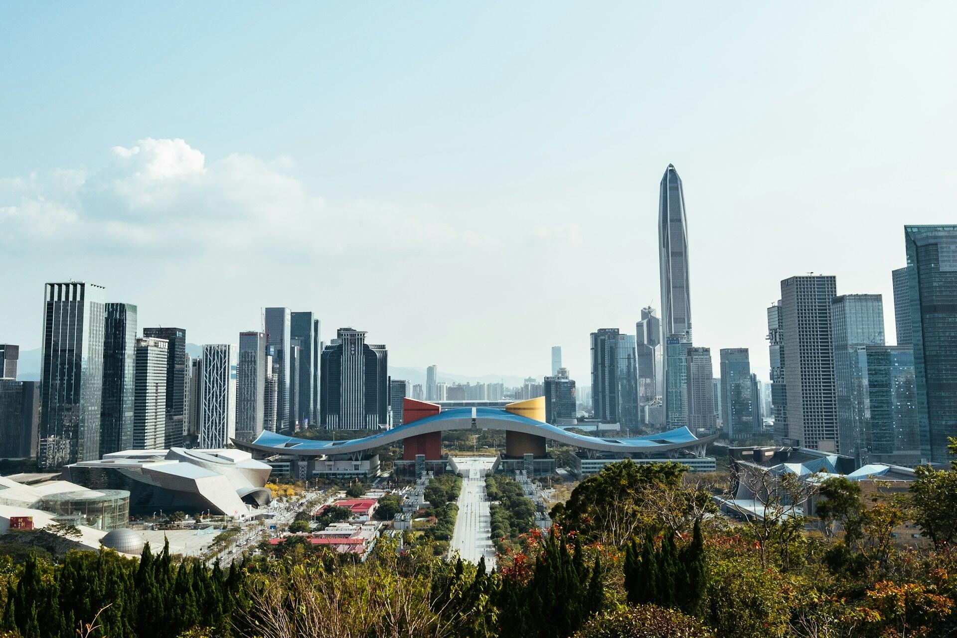 A panoramic view of a city focused on an ornate bridge spanning a walkway lined with greenery, and tall buildings in the background.