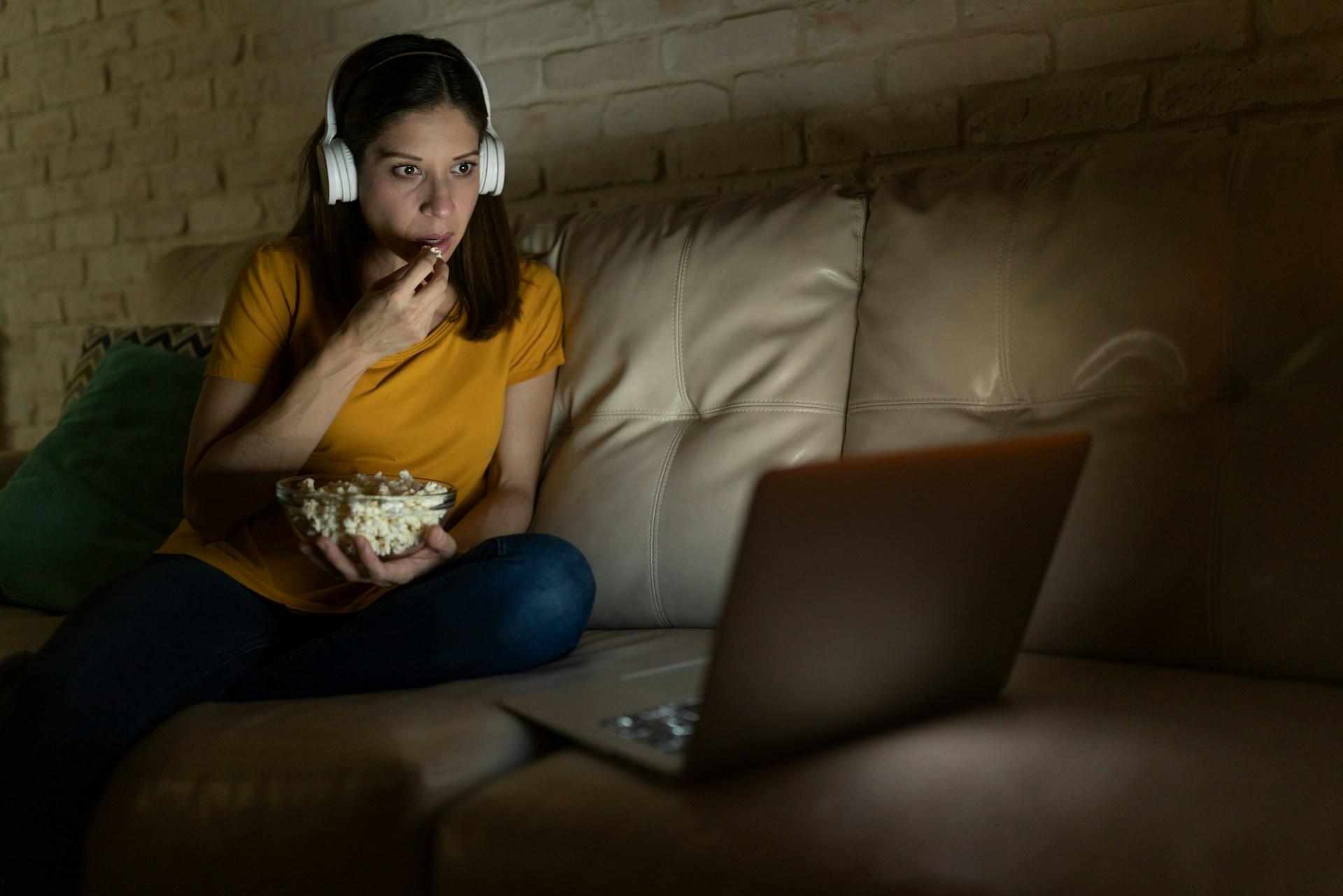 A person wearing an orange top and denims sits on a white leather couch with a glass bowl full of popcorn. They have white headphones on and watch a movie on their laptop computer, which sits on the couch.