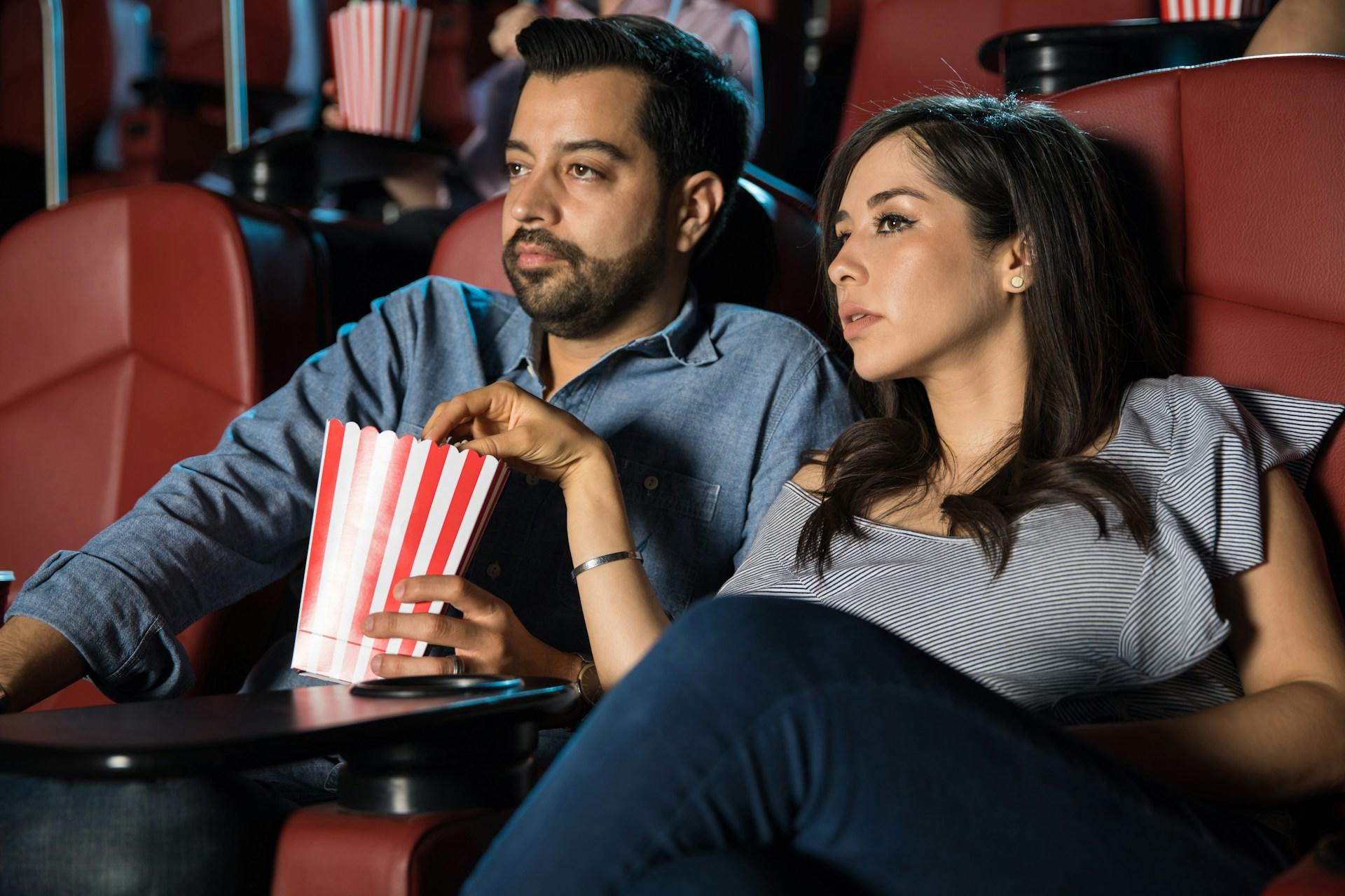A couple wearing casual clothing sits in red movie theatre chairs, sharing popcorn from a red and white striped bucket.