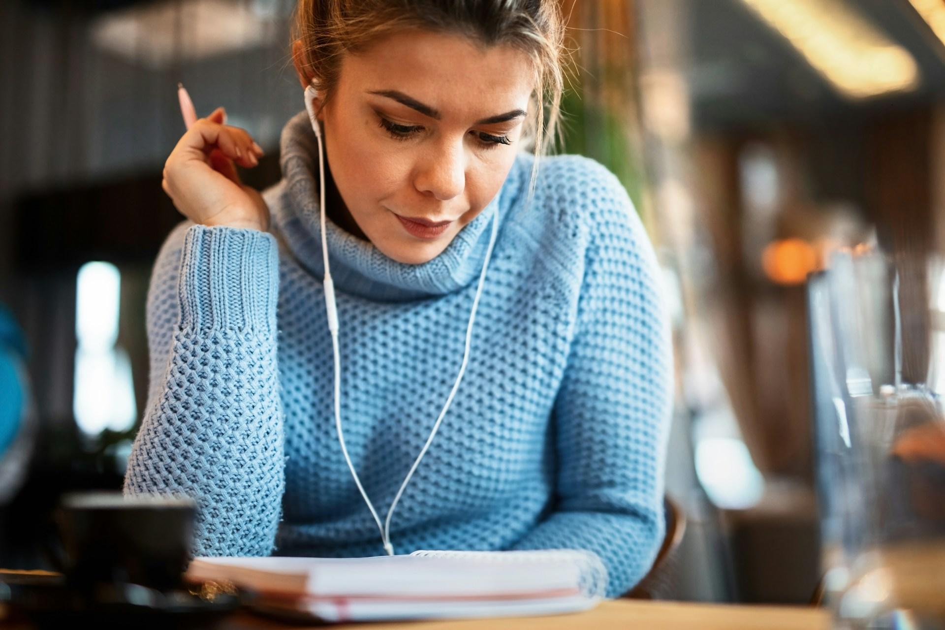 A person wearing a light blue jumper and earbuds with white cords in their ears sits at a wooden table with a spiral-bound notebook in front of them, looking downward.
