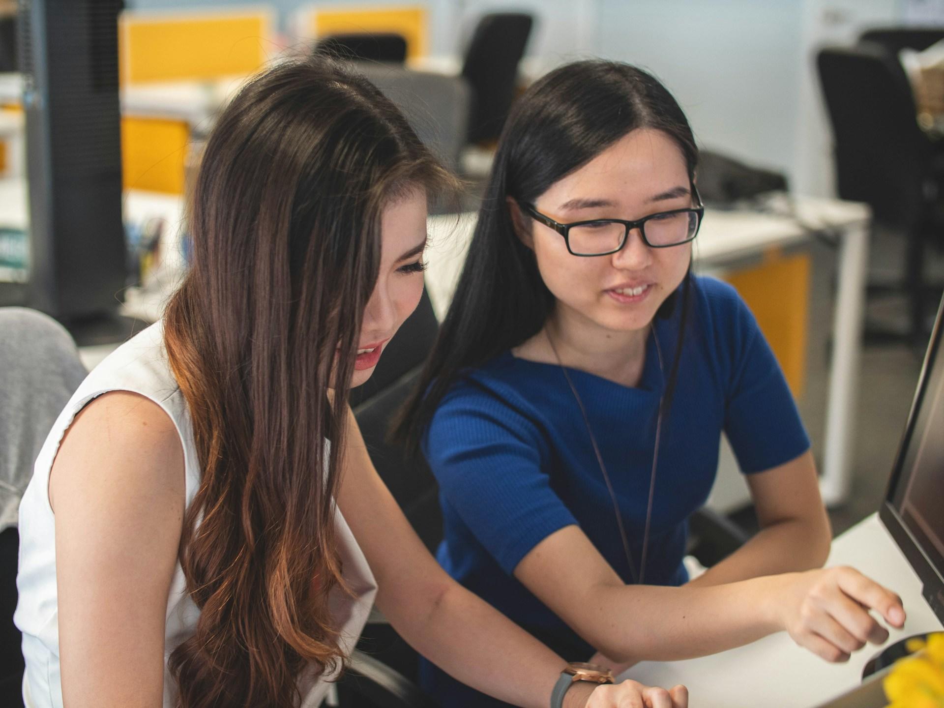 Two people sitting at a table. One wears a blue short-sleeved top and dark framed glasses and the other wears a white sleeveless top.