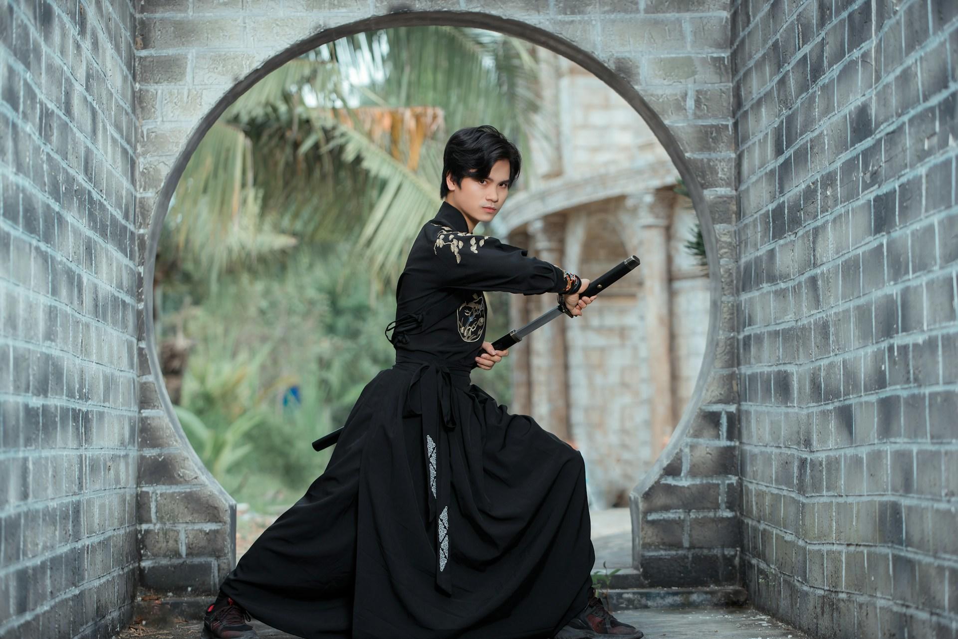 A young man with black hair is dressed in black samurai garb and prepares to draw his sword, posed in front of a stone arched gate between two grey stone walls.