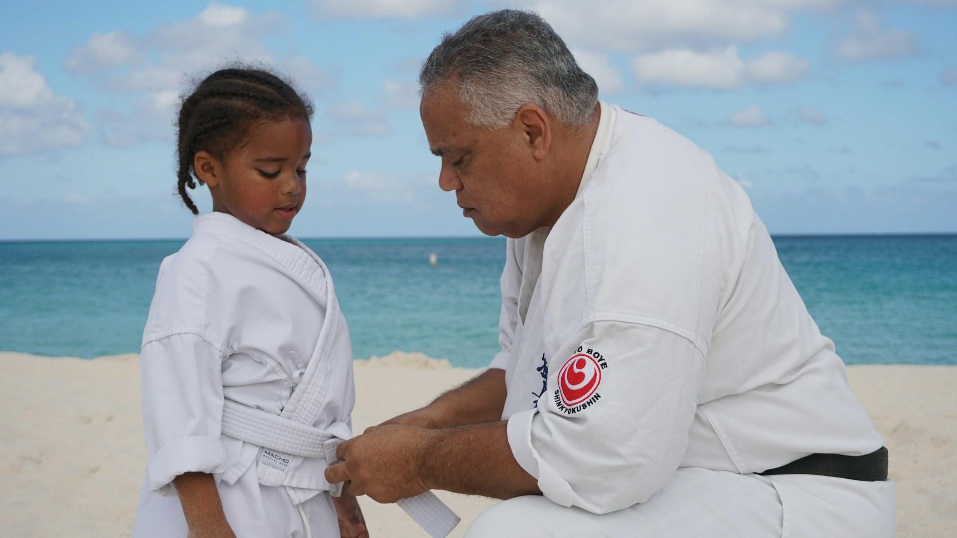 A sensei dressed in a white gi with black belt ties a student's white belt as they stand on the beach with the sea behind them.