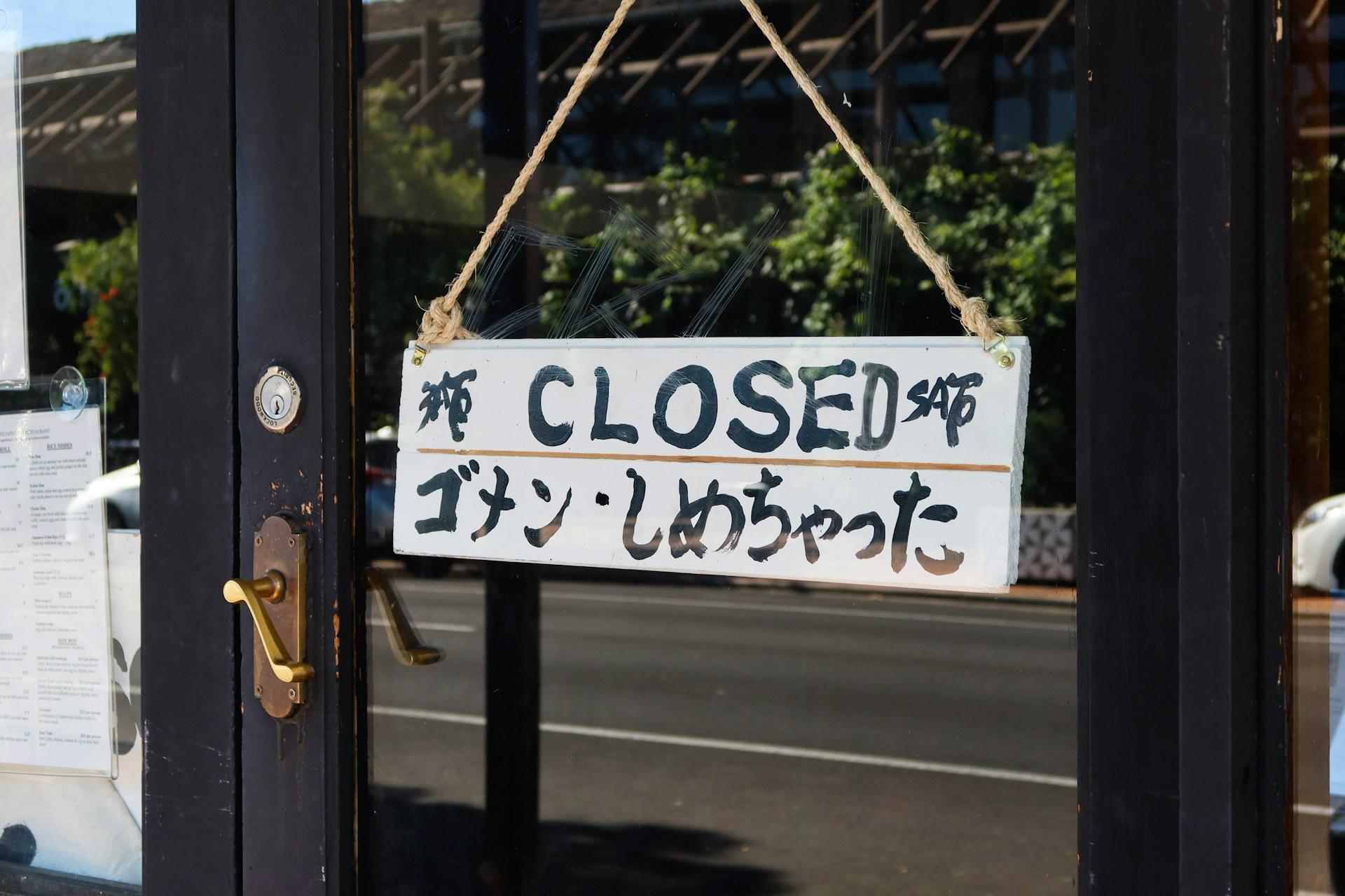 A shop sign with black letters and figures painted on a white background that say 'closed' in English and Japanese writing underneath. 