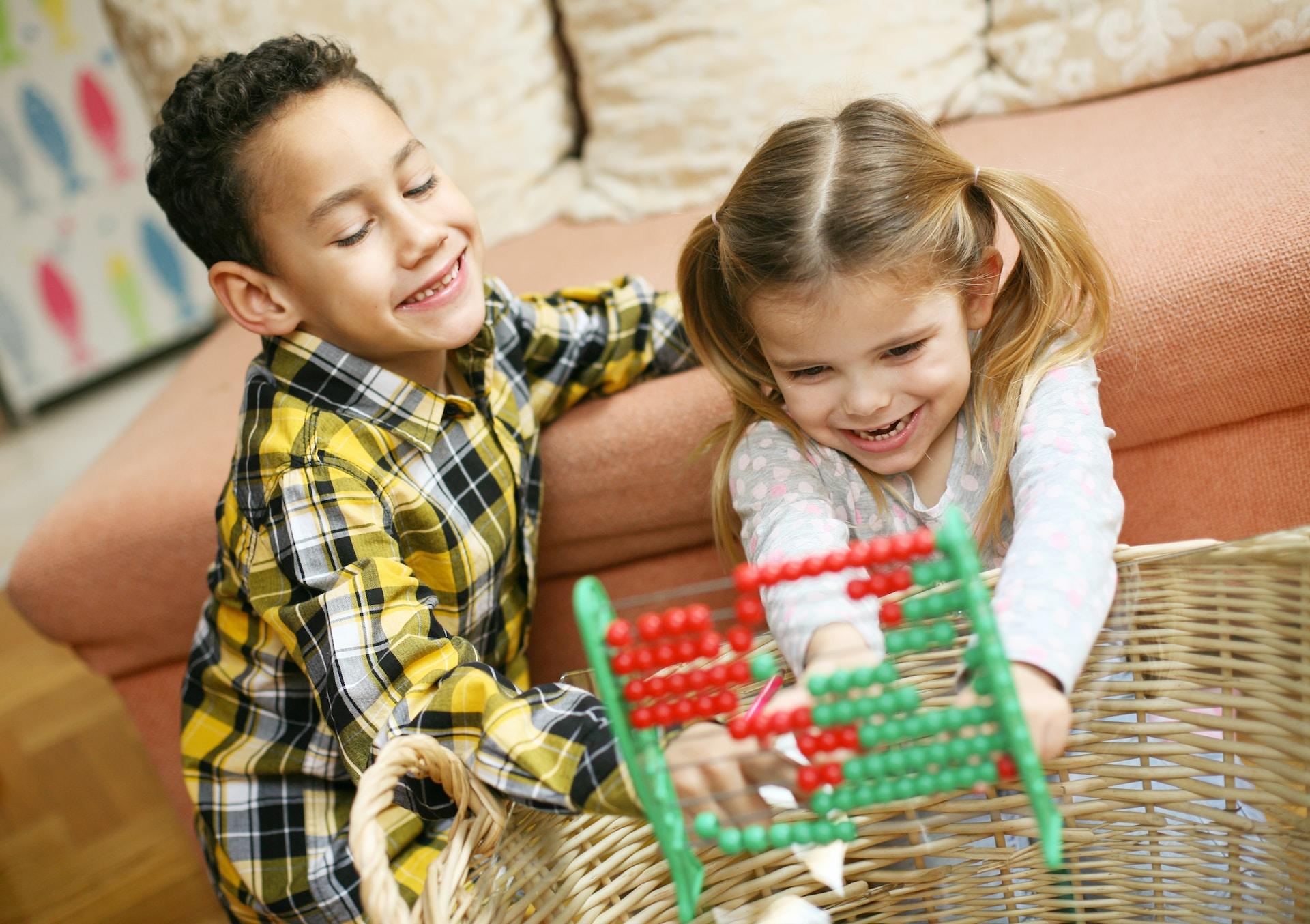 Two children smile as they kneel on the floor in front of a wicker basket holding a green and red abacus.