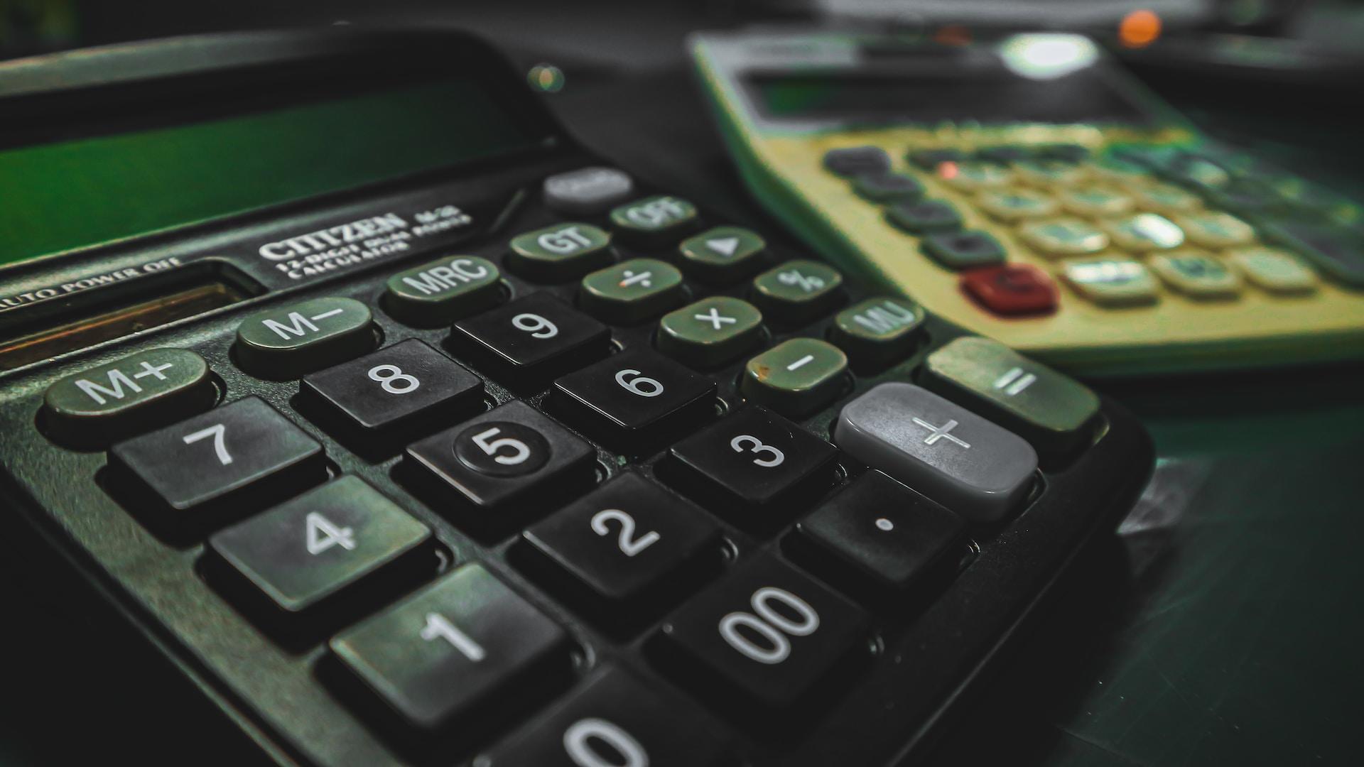 A close-up of a black pocket calculator with black number buttons, green function buttons and a grey equal button on a table with a white-faced calculator with red buttons in the background.