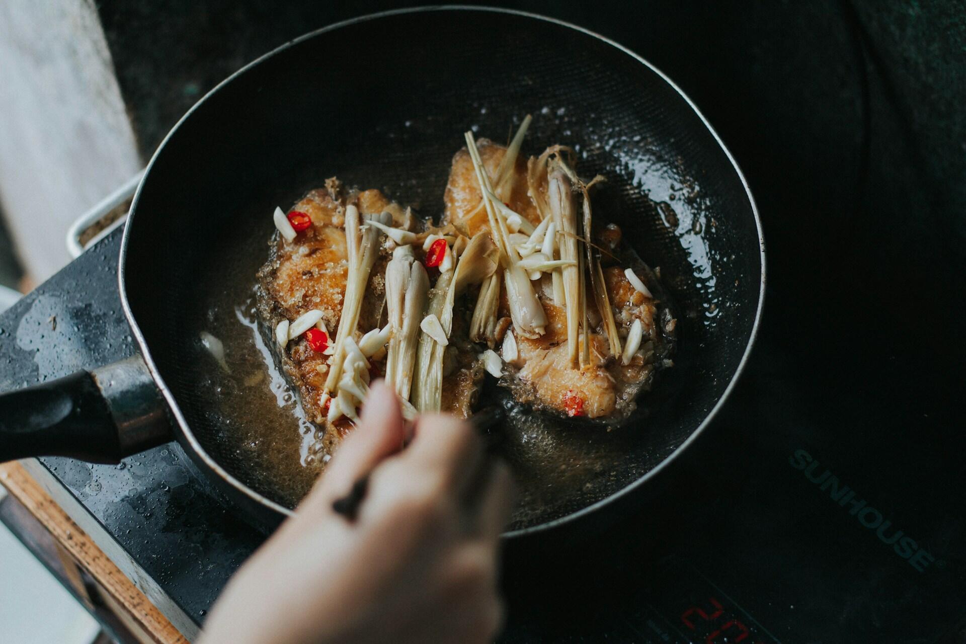 Fish frying in a wok with lemongrass and chili, hand stirring.