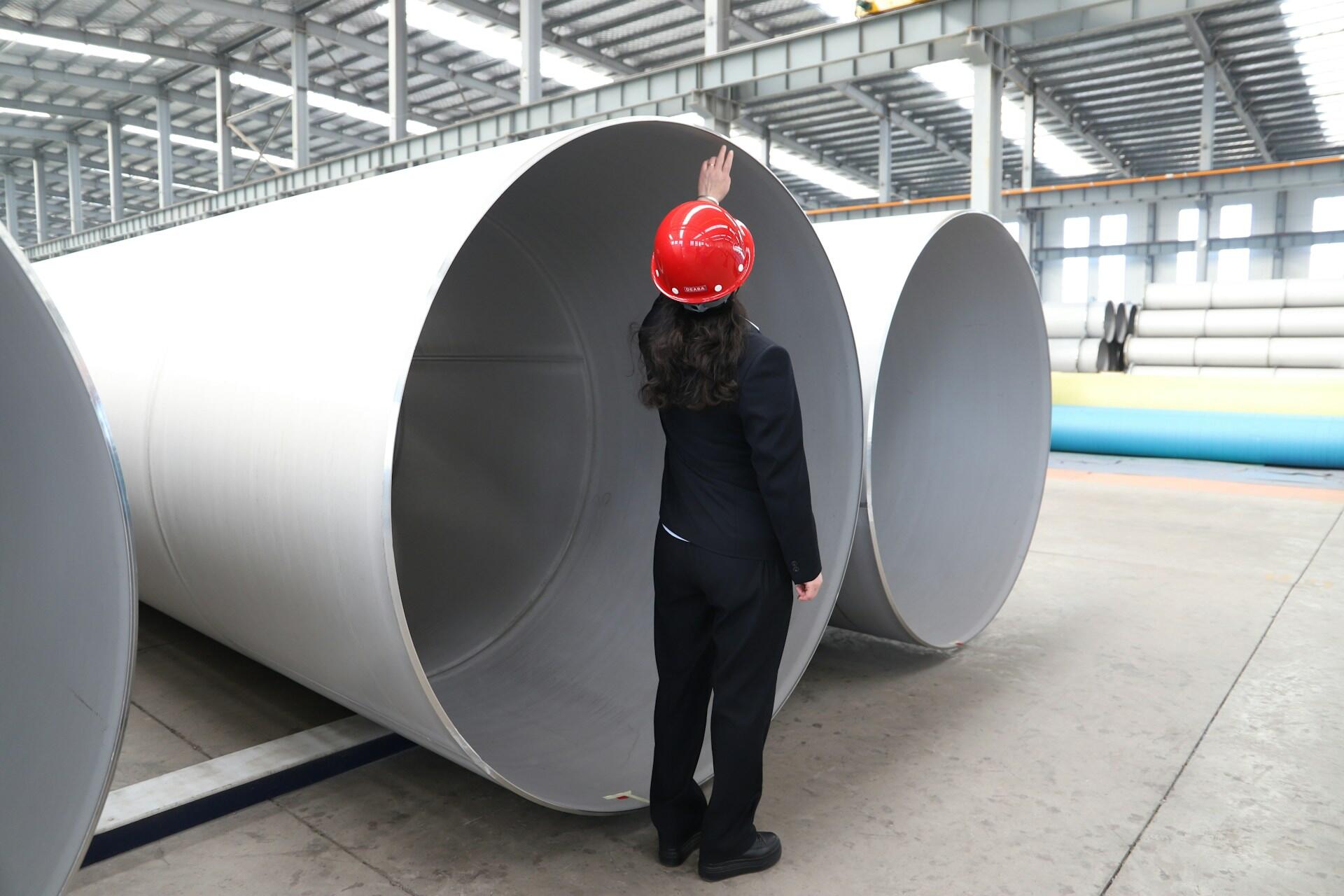 A person wearing blue coveralls and a red hardhat stands in front of a large pipe opening with their hand raised to the top edge.