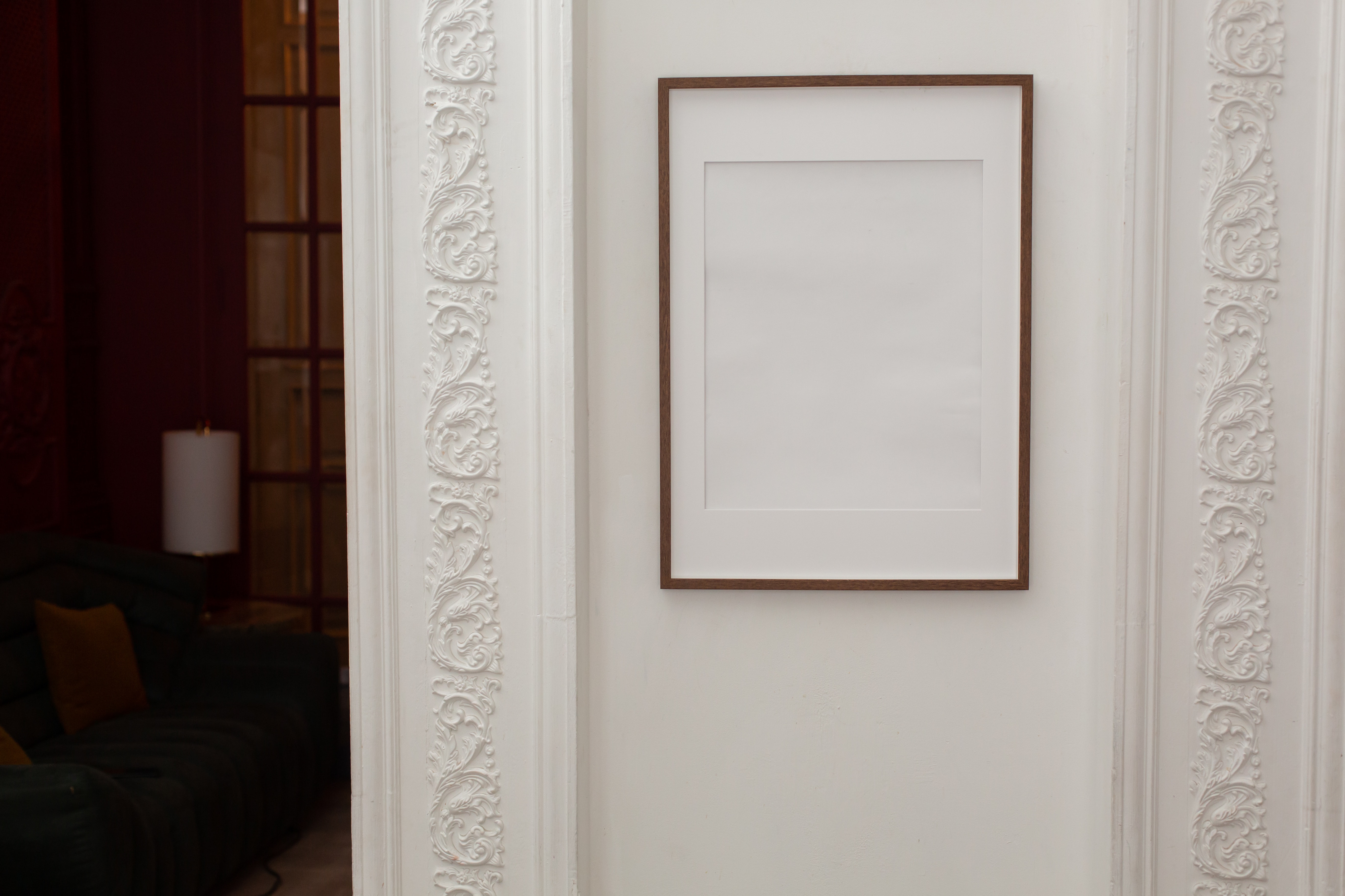 A brown, slim-line wooden portrait frame with a white mat but no picture hangs on a white wall trimmed with patterned edges. 