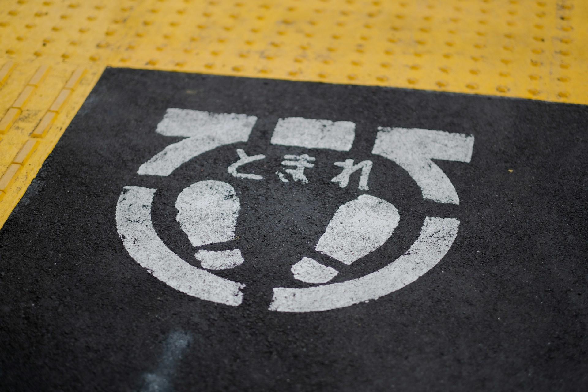 A stenciled street sign on the pavement, painted with white paint against a dark background, shows Japanese characters atop a set of footprints.