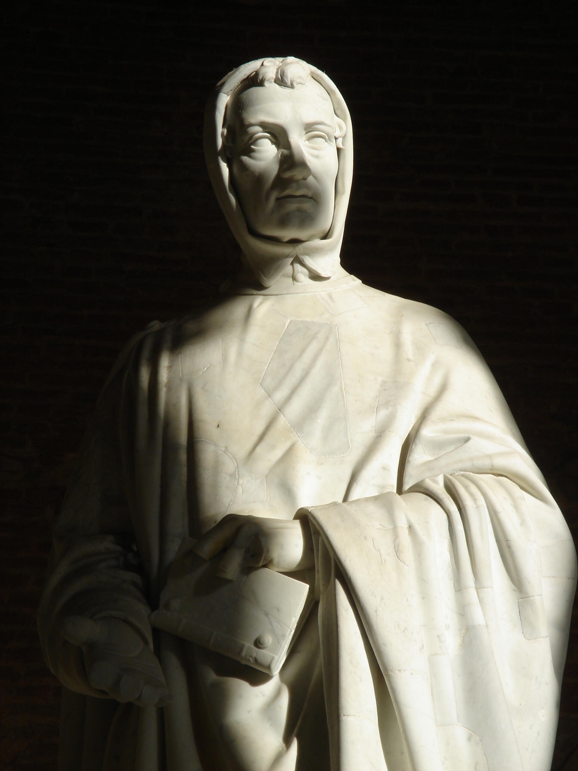 A marble statue of a robed, hooded person holding a book against a black background.