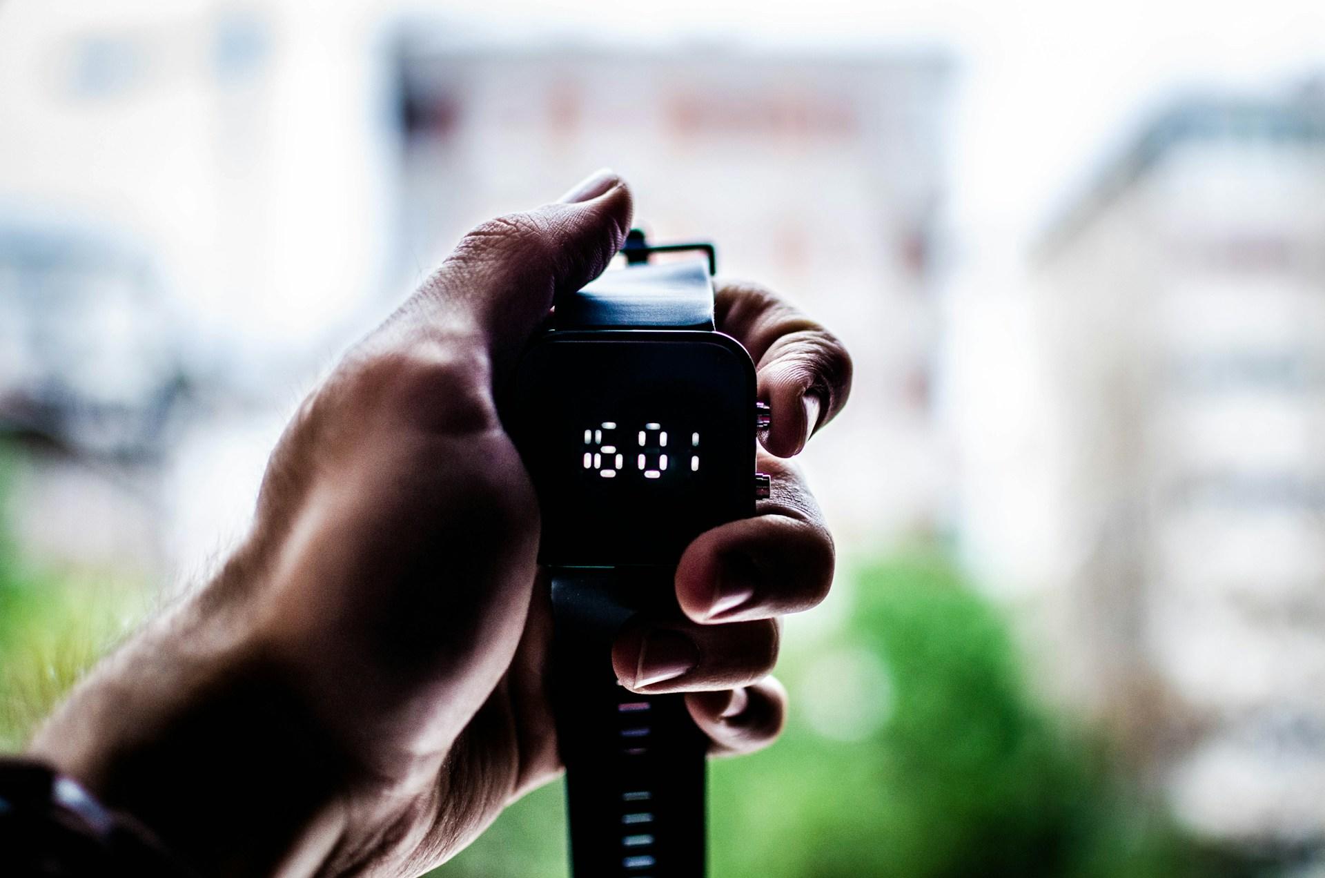A person holds a digital stopwatch with a black case displaying the number 16:01 against a blurred background. 