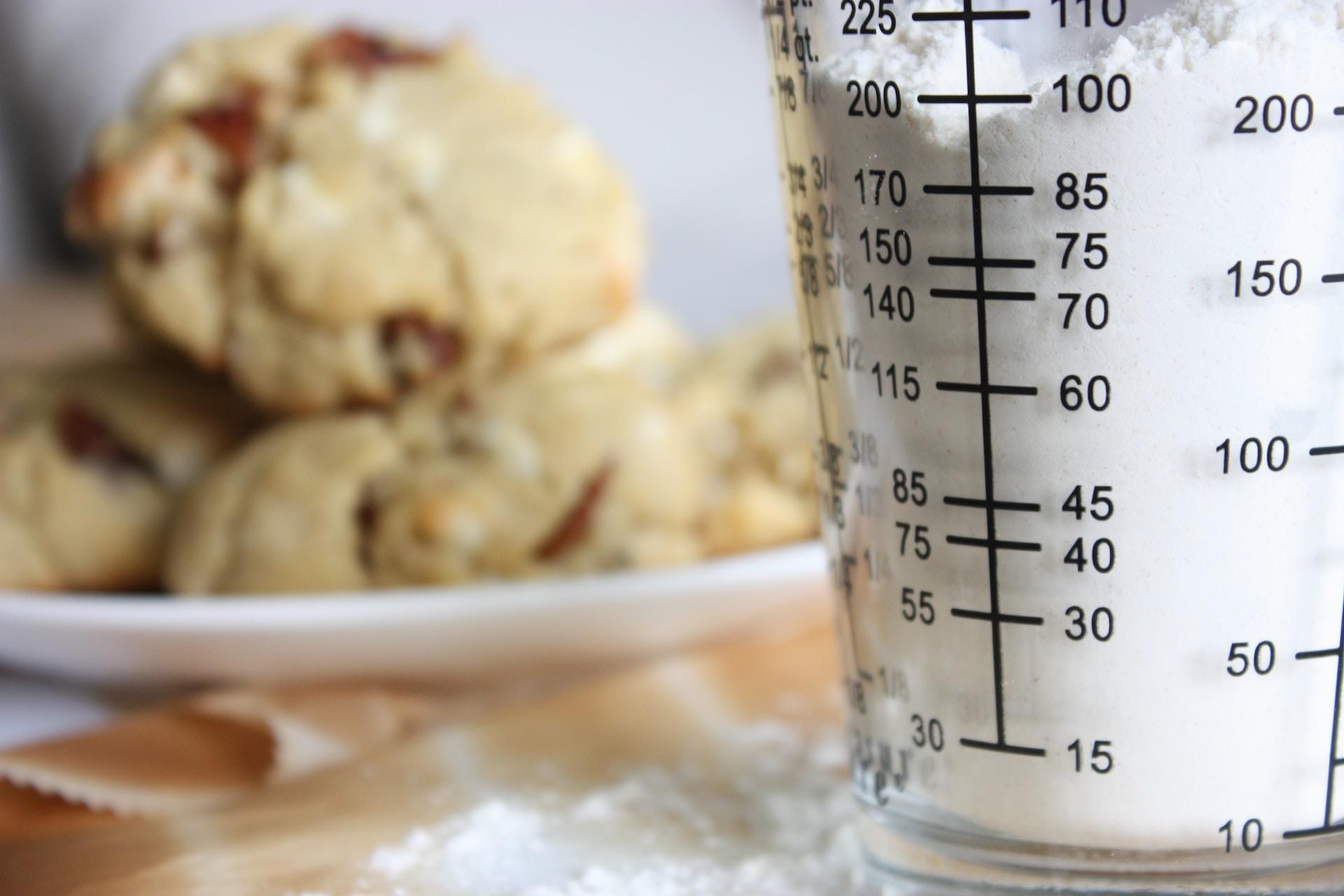 A clear measuring cups with black graduated markings is filled with flour. A plate of scones is blurry in the background.