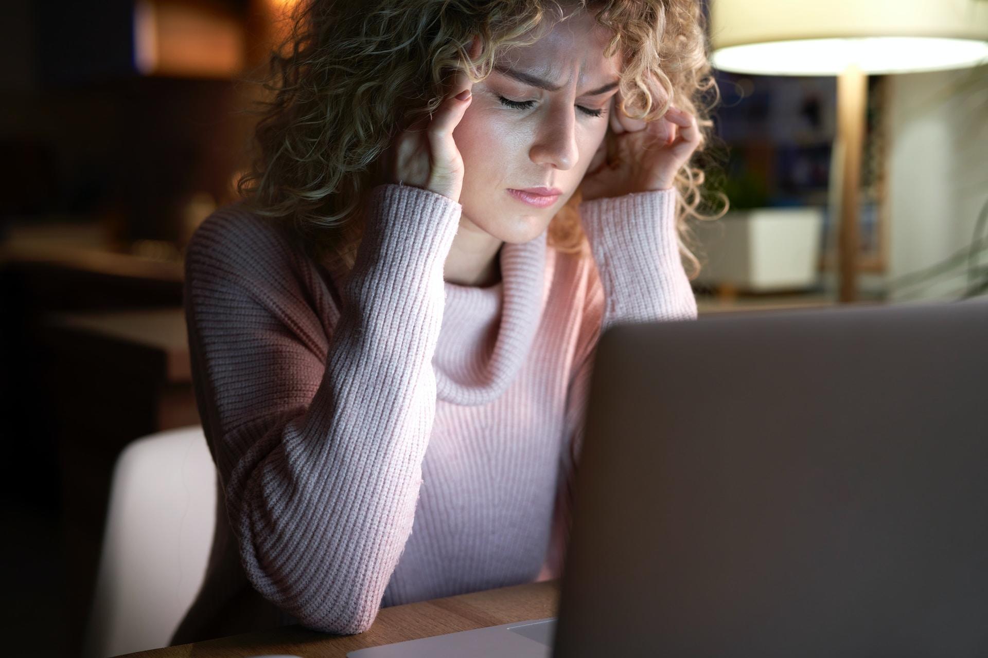 A person wearing a white shirt sits in a darkened room in front of an open laptop, which casts its glow on your distressed face and their hands pressing on their temples.