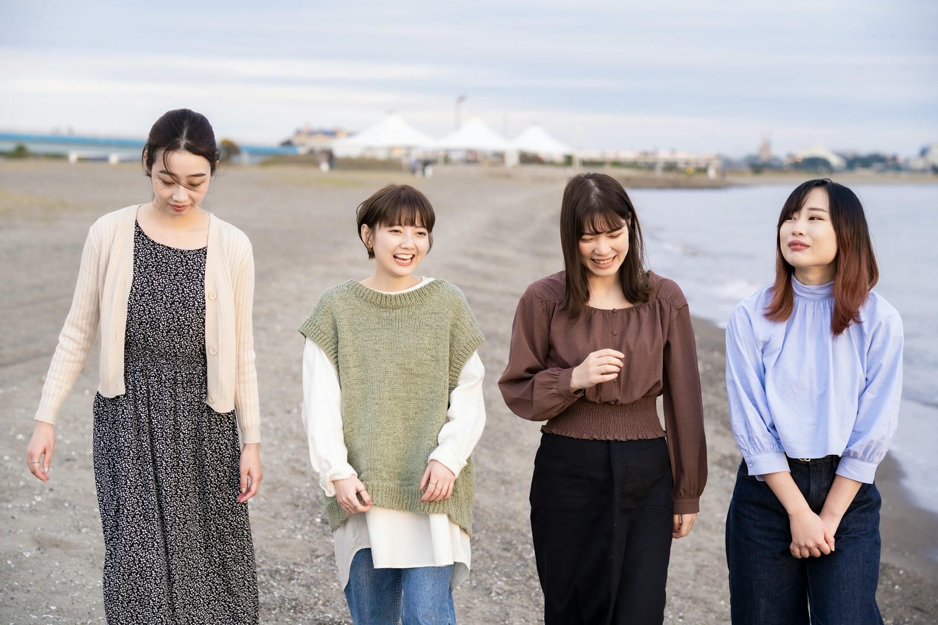 Four women with Asian appearance walk together on the beach, smiling and talking. 