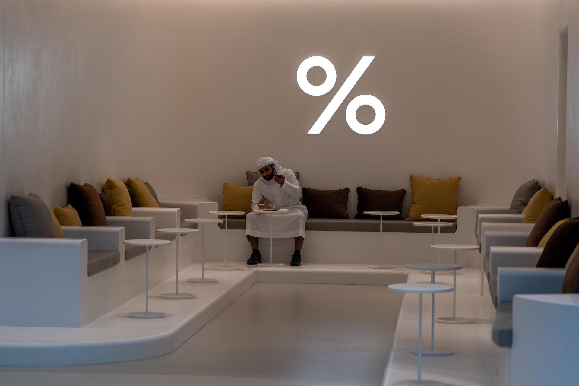 A Dubai airport lounge, empty save for one person dressed in white in an otherwise white room with muted brown tones and a large percentage sign lit up in white neon.