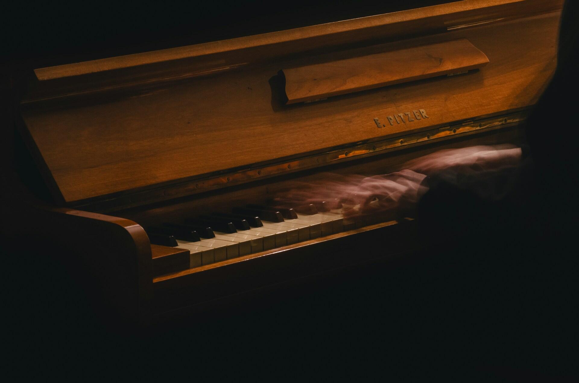 A blurred hand playing a wooden piano's keys, capturing the motion and artistry of live music in a dimly lit setting.