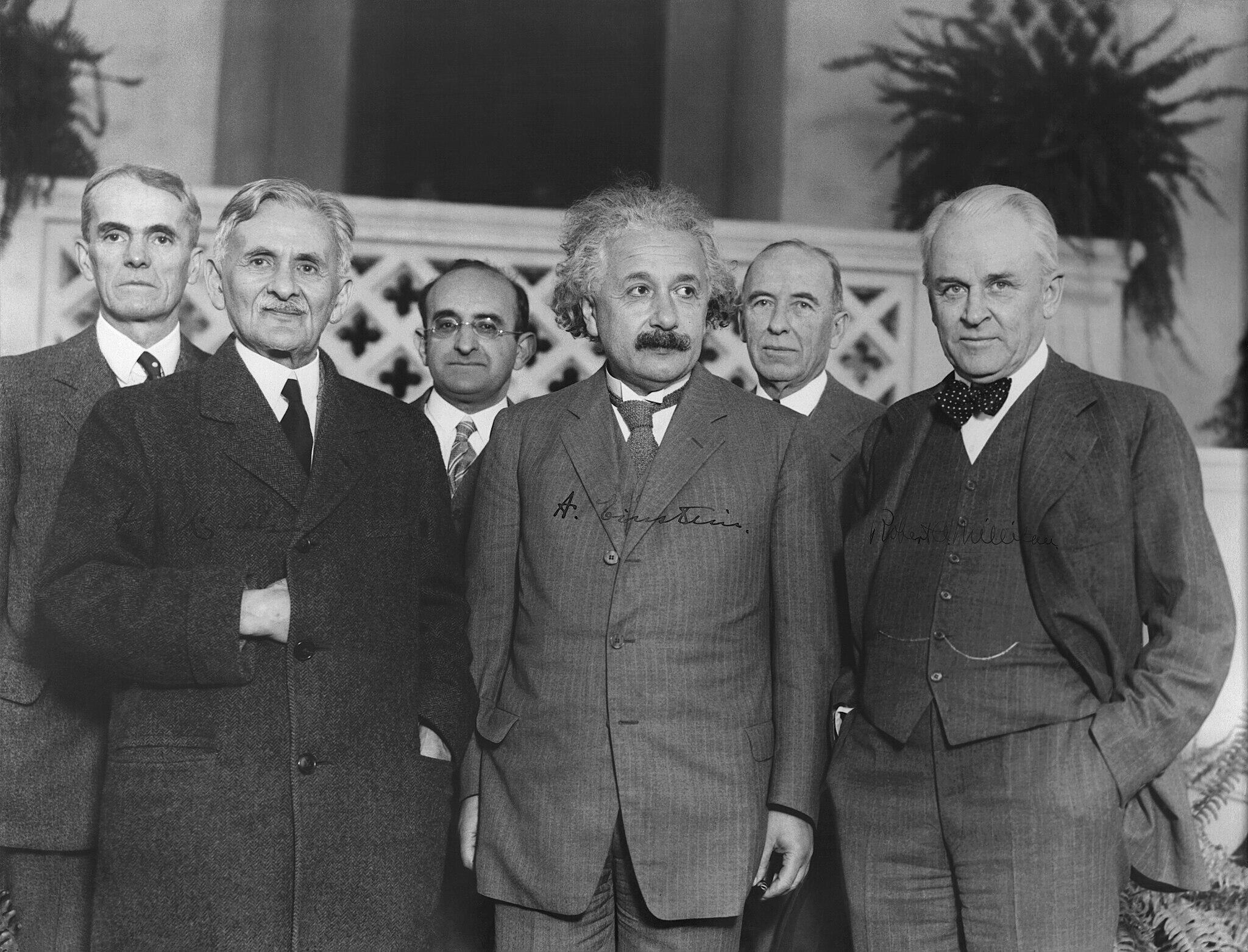 Black-and-white photo of seven men, including Albert Einstein, in suits standing indoors, with decorative plants in the background.