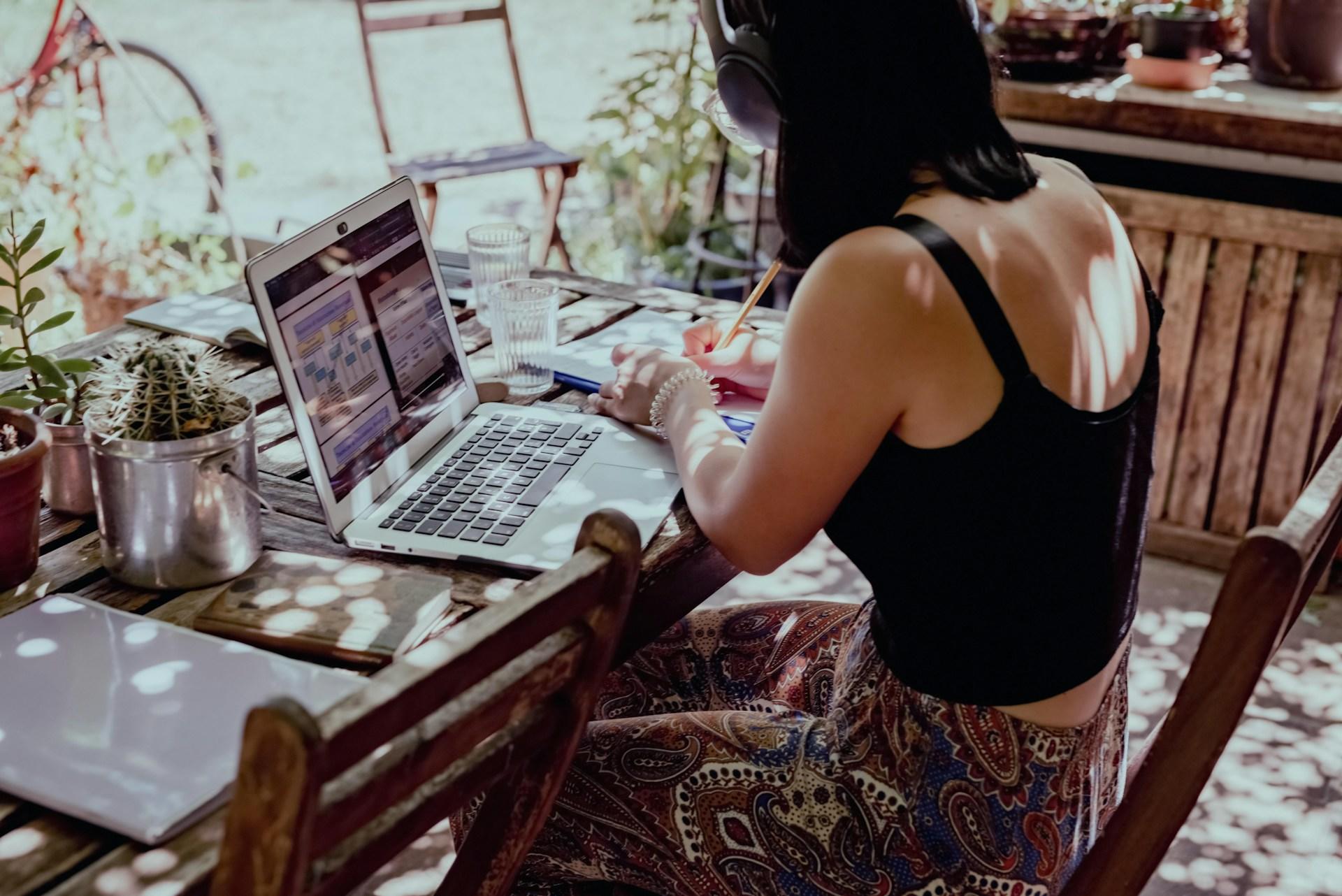 A person wearing a dark coloured tank top sits at a wooden table that has an open laptop computer on it, writing notes.