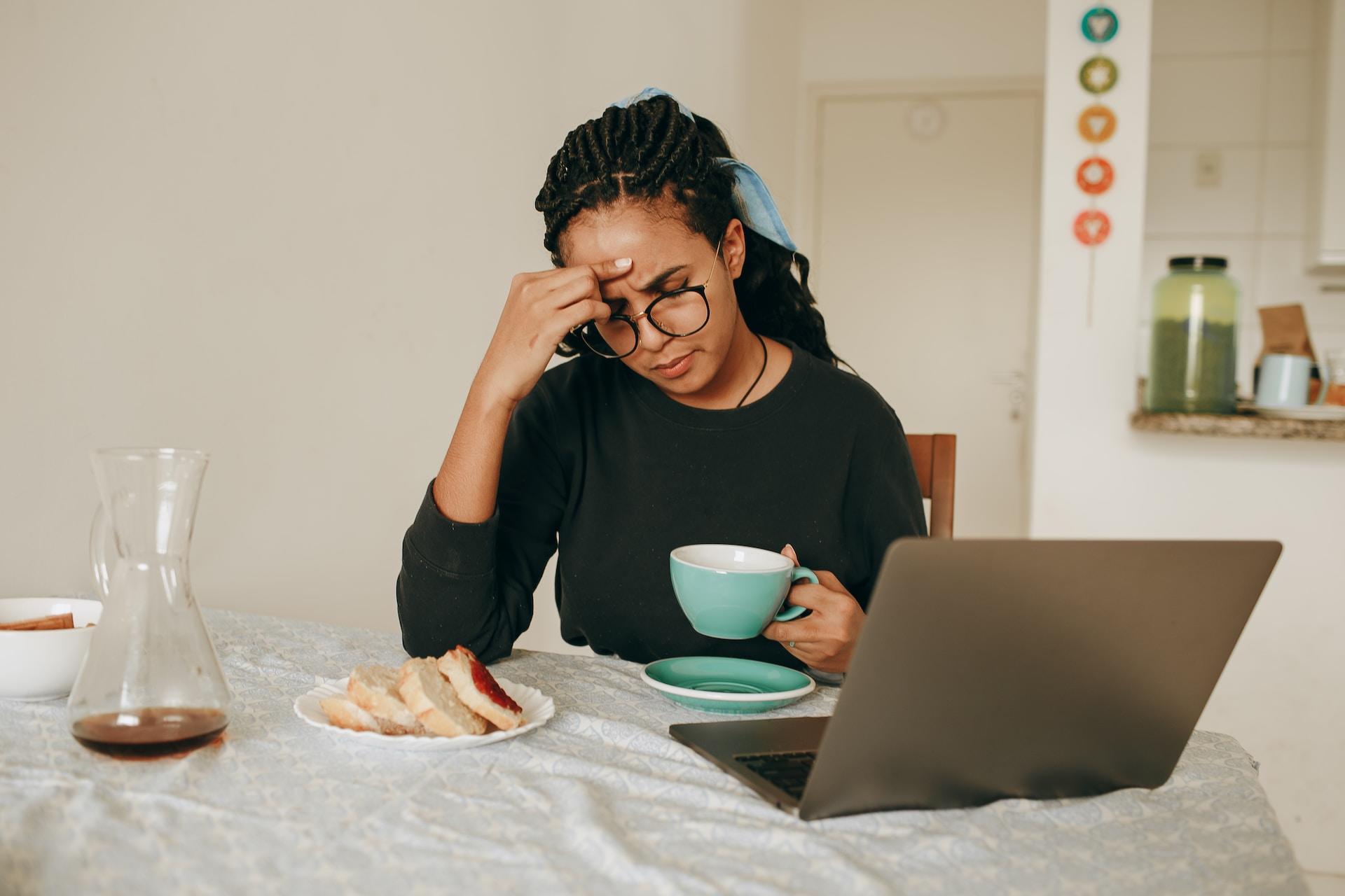 A person wearing a black top holds a green cup of drink while sitting at a white table with an open laptop in front of them and a plate of sweet treats to one side.