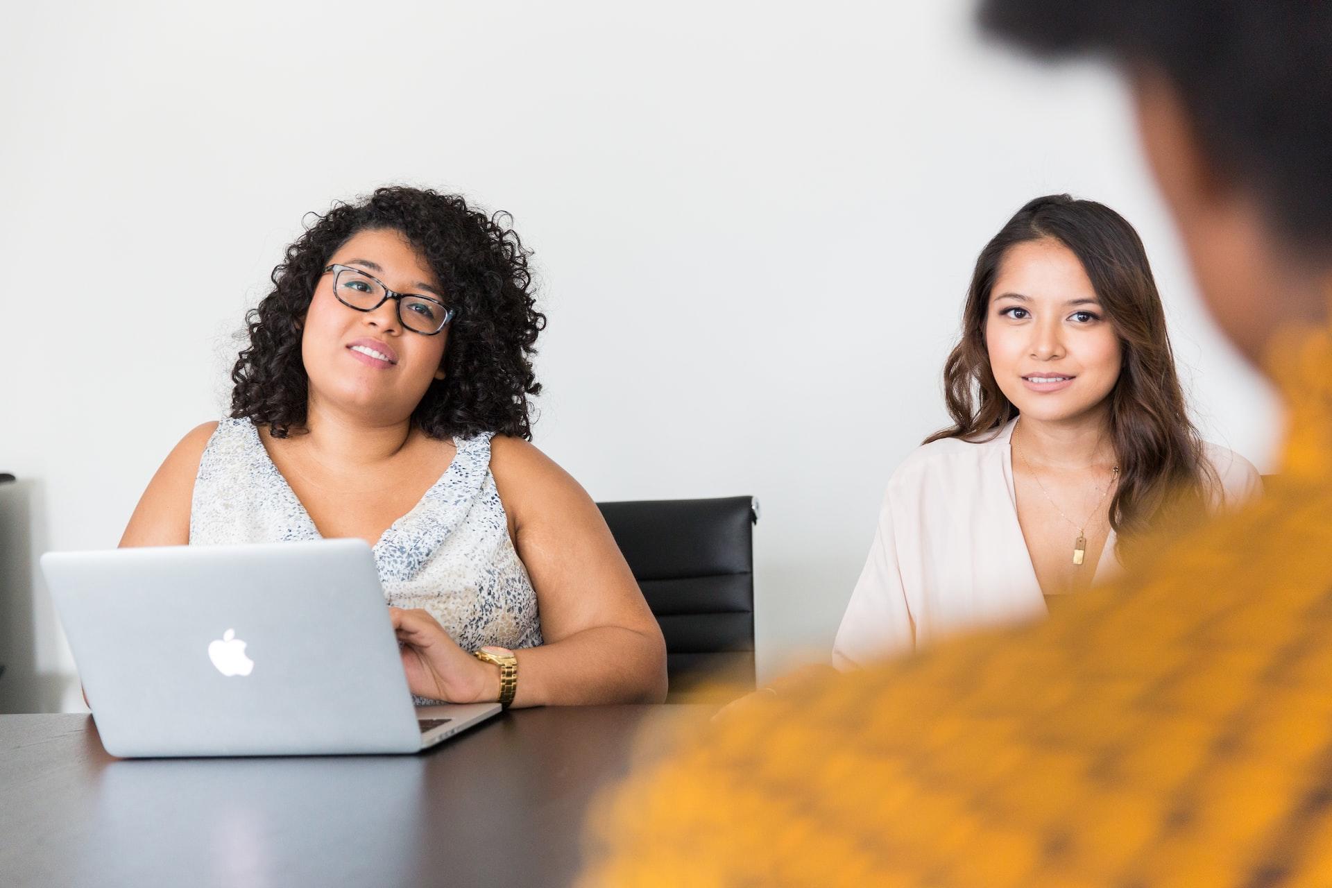 One woman wearing glasses and a sleeveless top sits in front of an open laptop computer next to another another woman in a white top. They are across the table from a person wearing a yellow and black patterned shirt. 