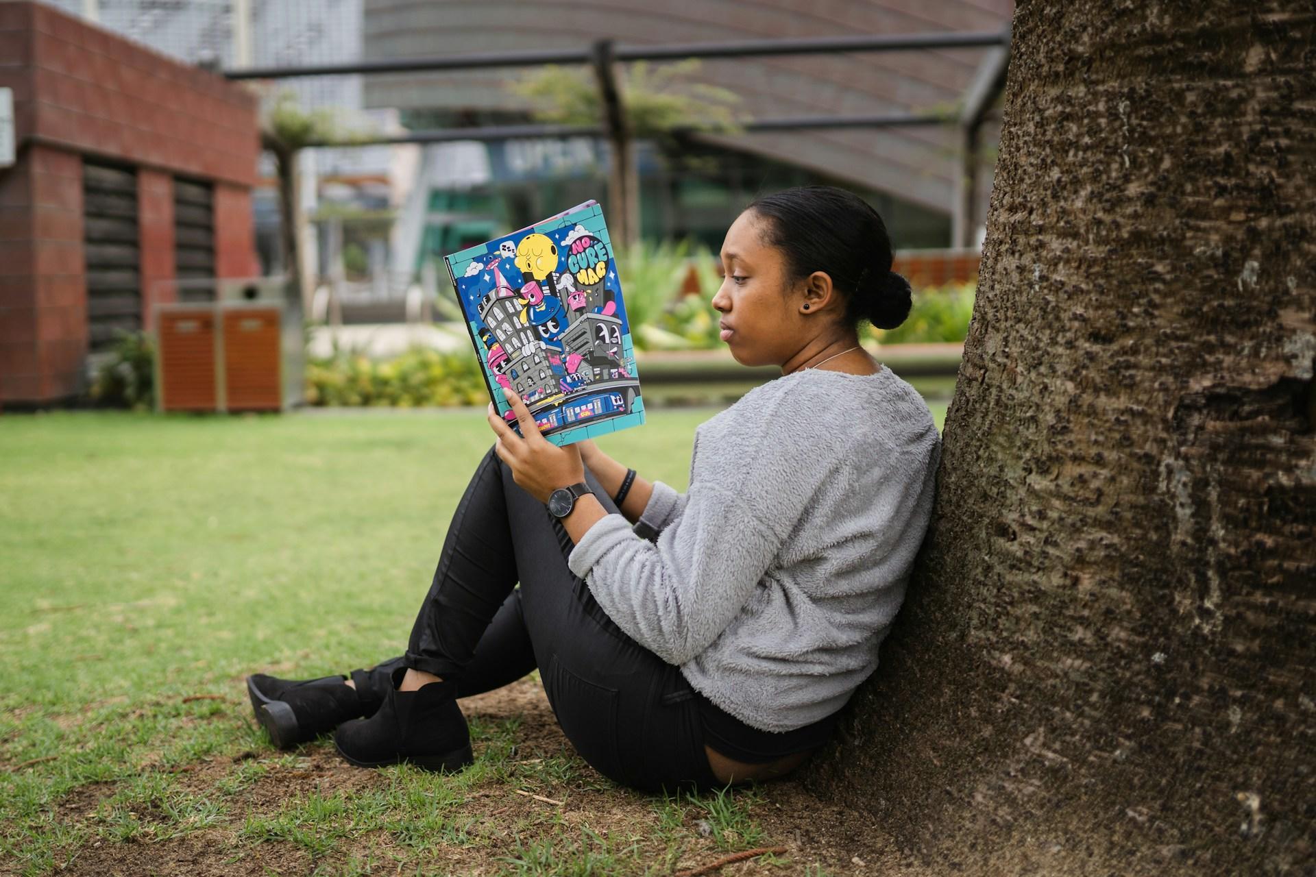 A person wearing a grey top and black trousers sits on the green grass, propped against a tree, reading a book lively-coloured cover.