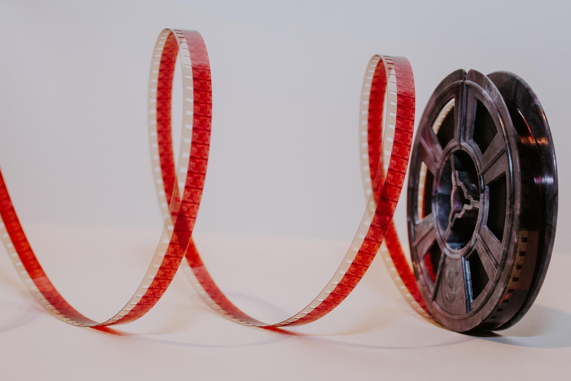 A grey film reel seen against a neutral background with a string of red film spooling off of it. 