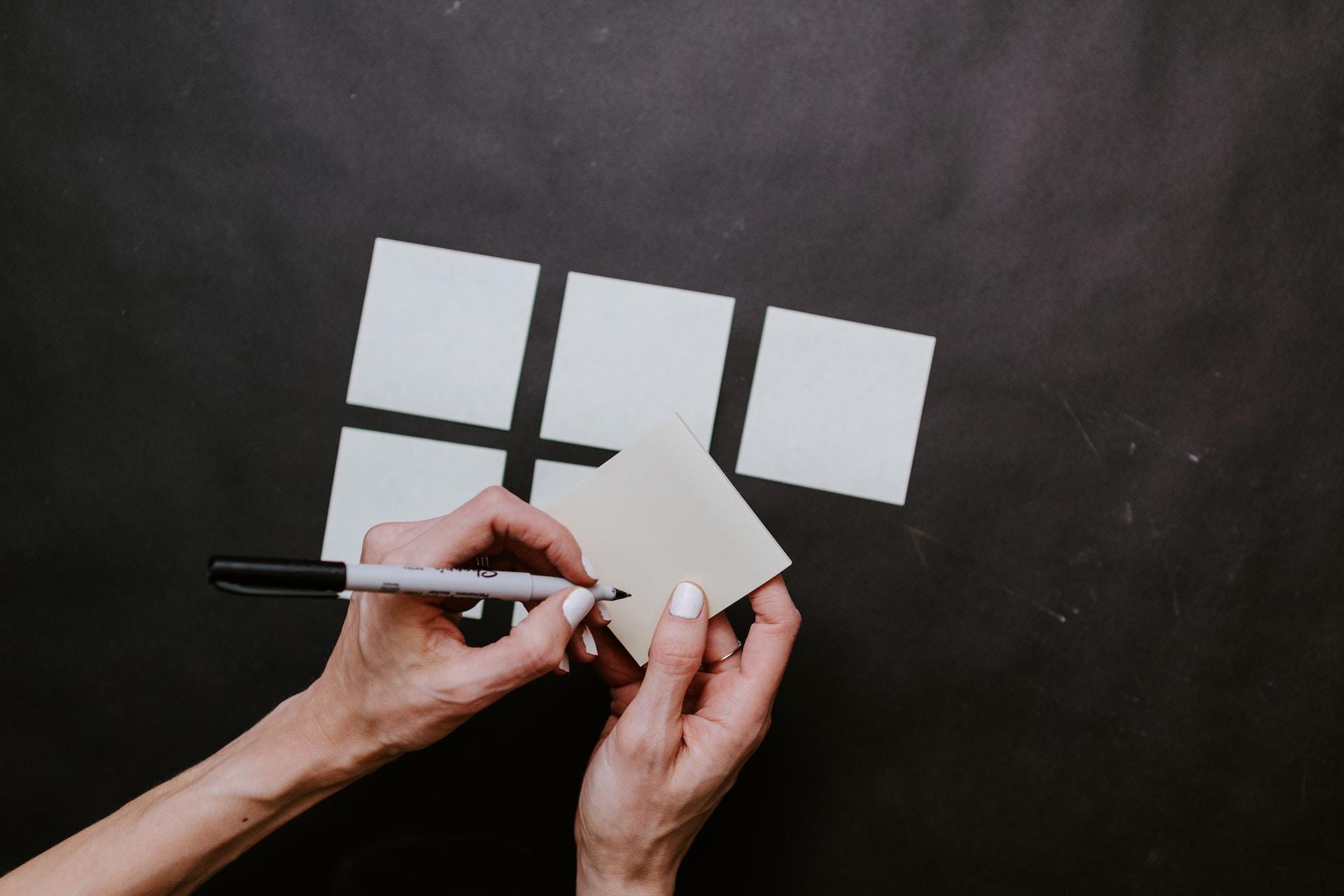 Six squares of white paper lay against a black surface. A person holds the bottom right square in their right hand, preparing to write on it with a black Sharpie marker. 