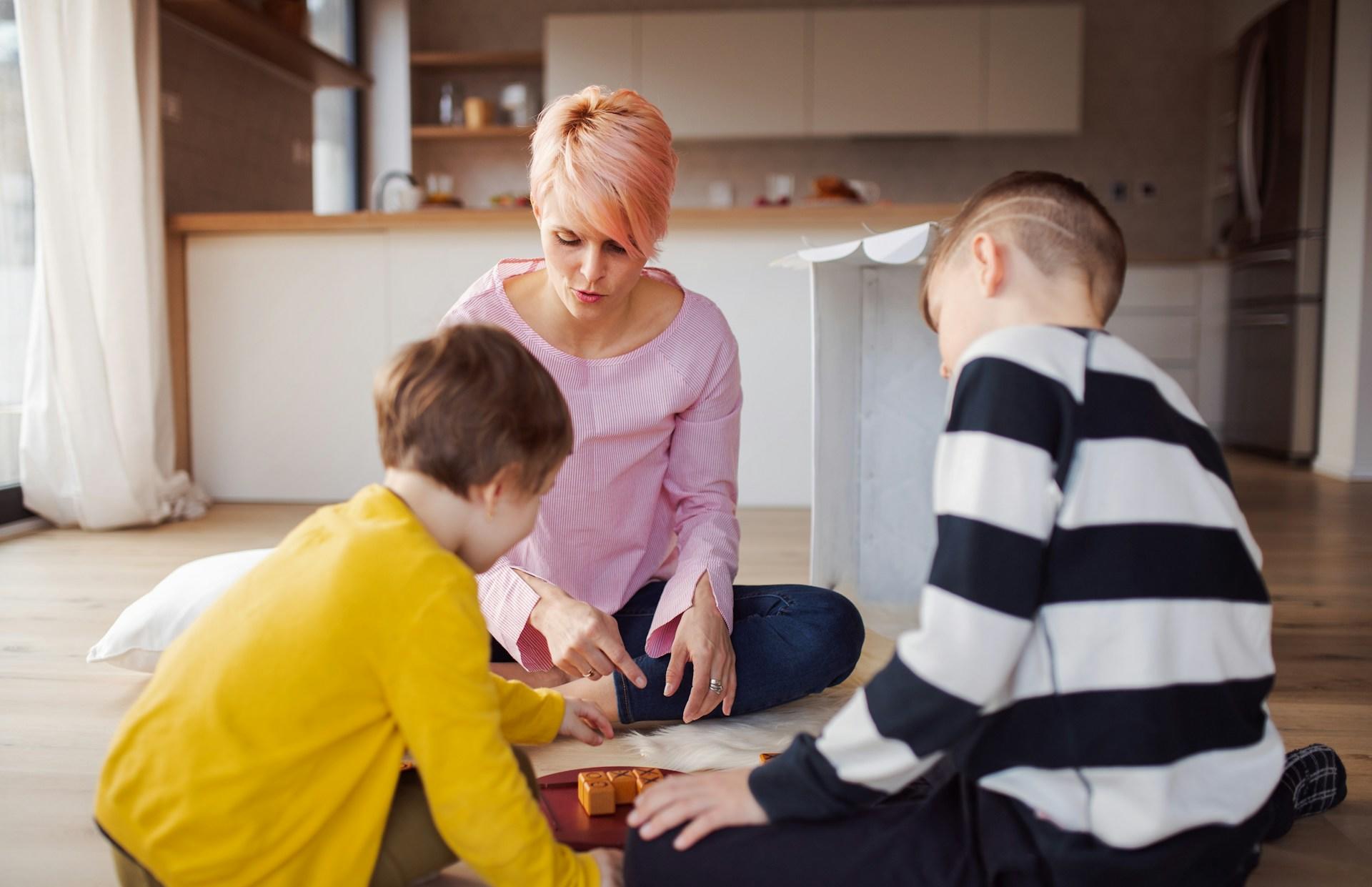 A youth wearing a shirt with black and white horizontal stripes sits next to a younger child wearing bright yellow. They both sit opposite an adult wearing pink with pink-toned hair, playing a game on the floor, in front of a kitchen setup. 