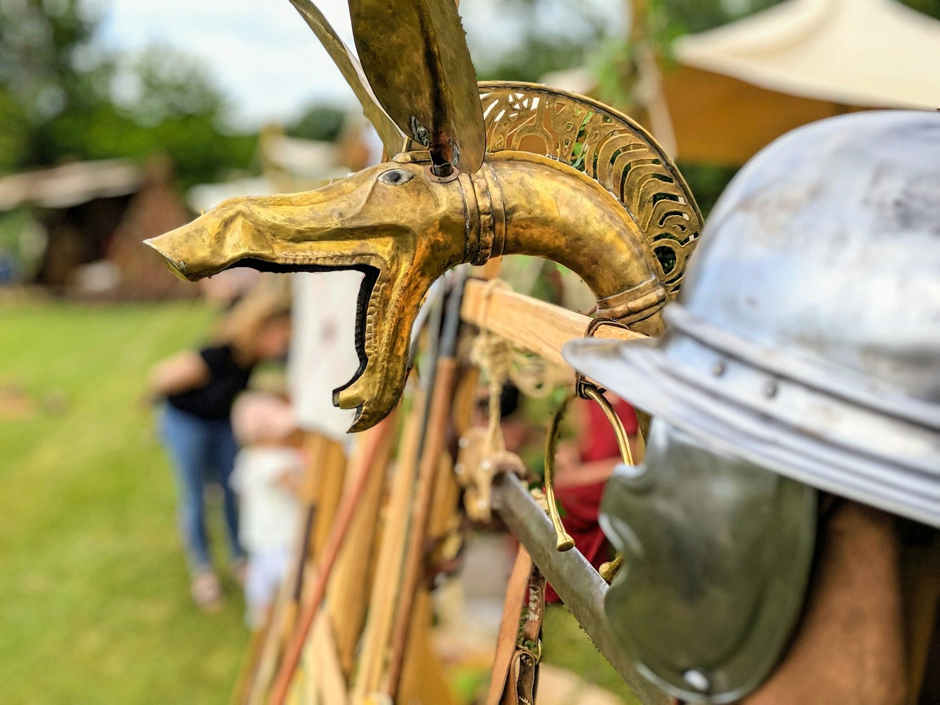 A brass dragon movie prop with its mouth wide open waits for its appearance next to a silver roman cask and other props on an outdoor set in an expansive green meadow.