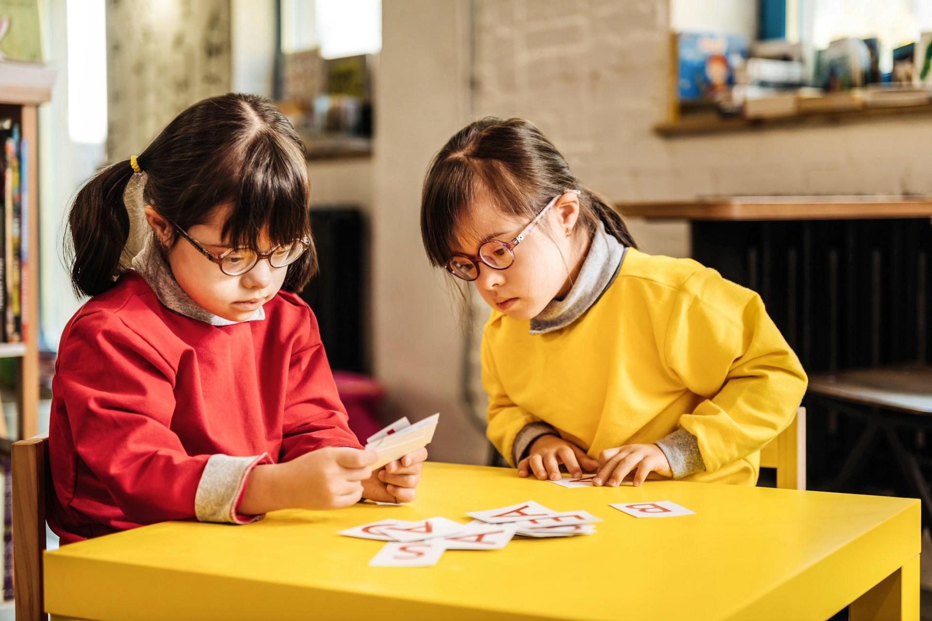 Two small girls wearing dark framed glasses, one in a red shirt and the other wearing yellow, sit at a bright yellow table looking at game cards. 