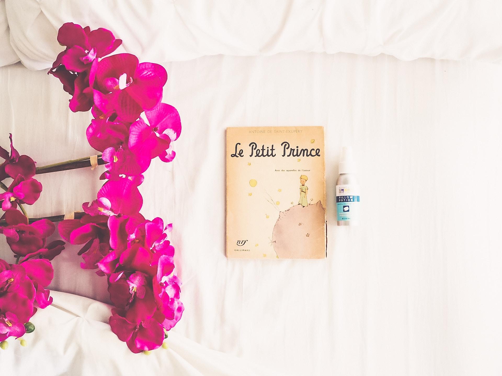 A copy of The Little Prince laying next to fuschia coloured flowers on a white linen surface.