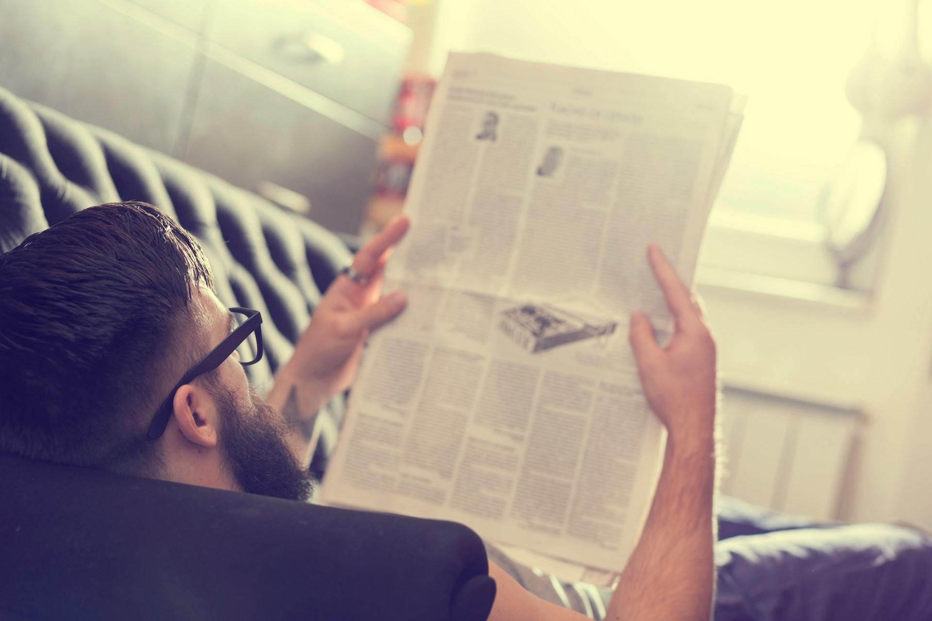 A person wearing dark-framed glasses and dark clothing reclines in a white hammock, reading a newspaper.
