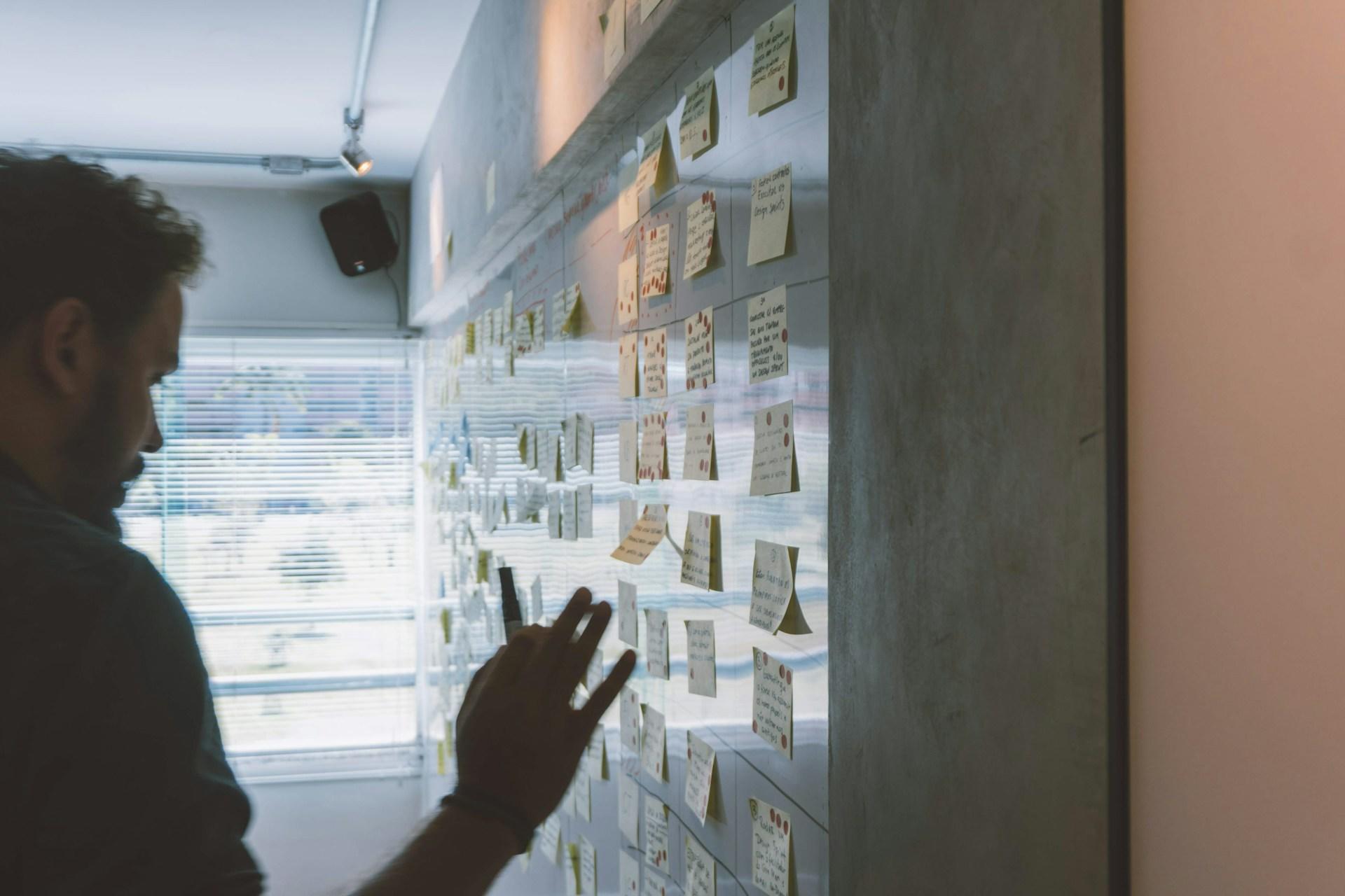 A person seen in silhouette in an indirectly-lit room stands before a board loaded with sticky notes, holding one in their hand. 