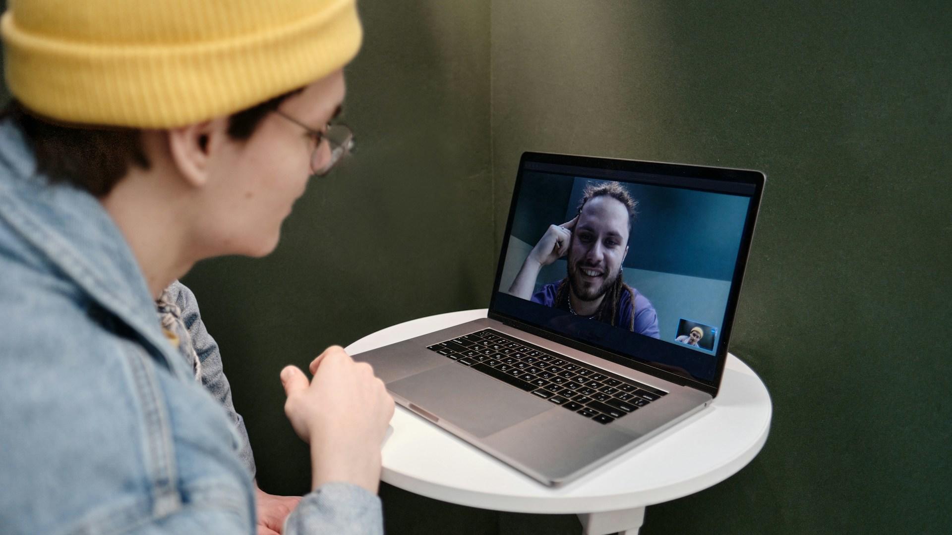 A person wearing a light blue long sleeved top and a yellow beanie sits in front of a silver laptop computer whose screen shows a smiling person with their head propped up on their hand.