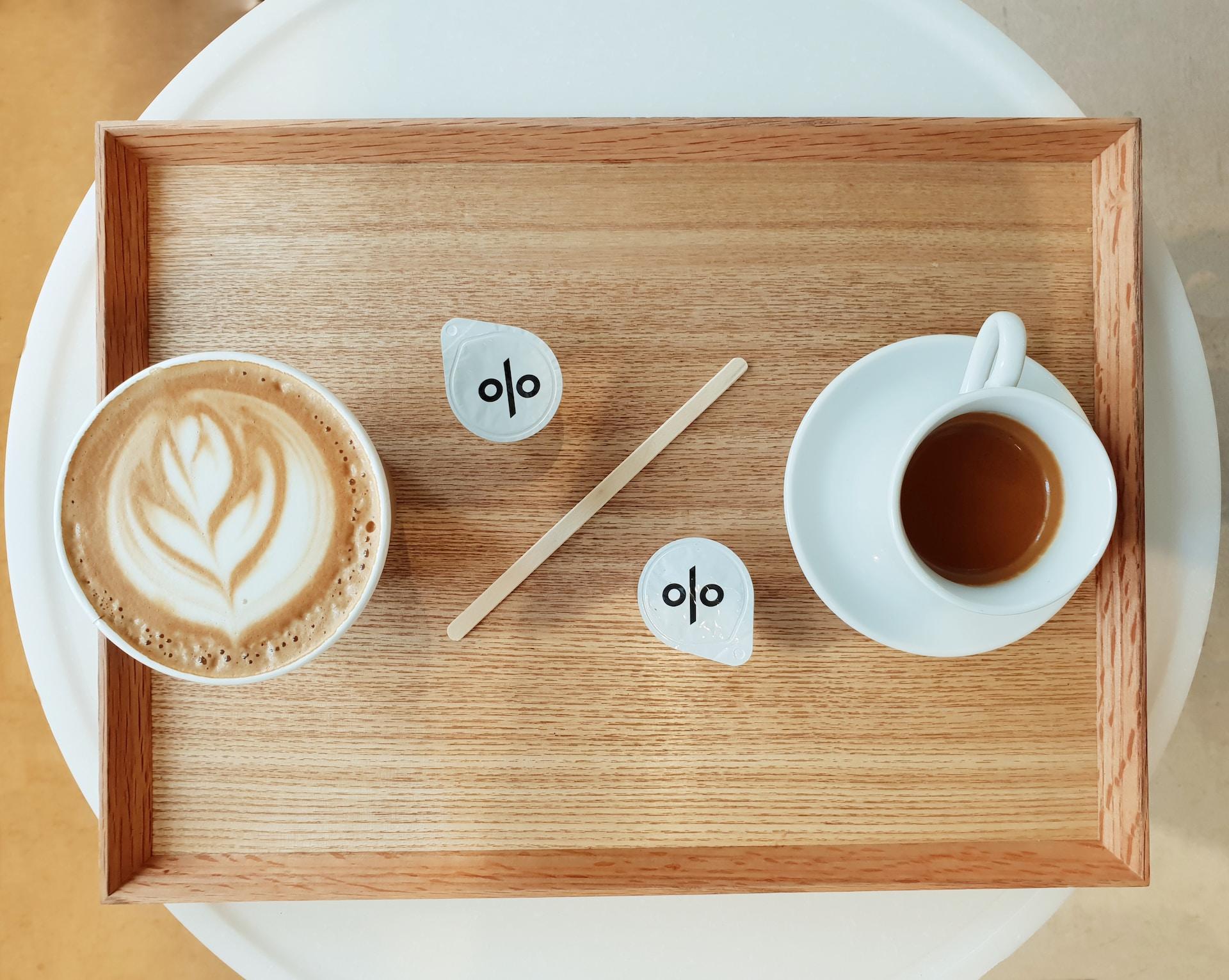 A bamboo tray on a white table with a coffee art latte and a cup of black tea on it, separated by two coffee milk pods and a stir stick which form a percentage sign.