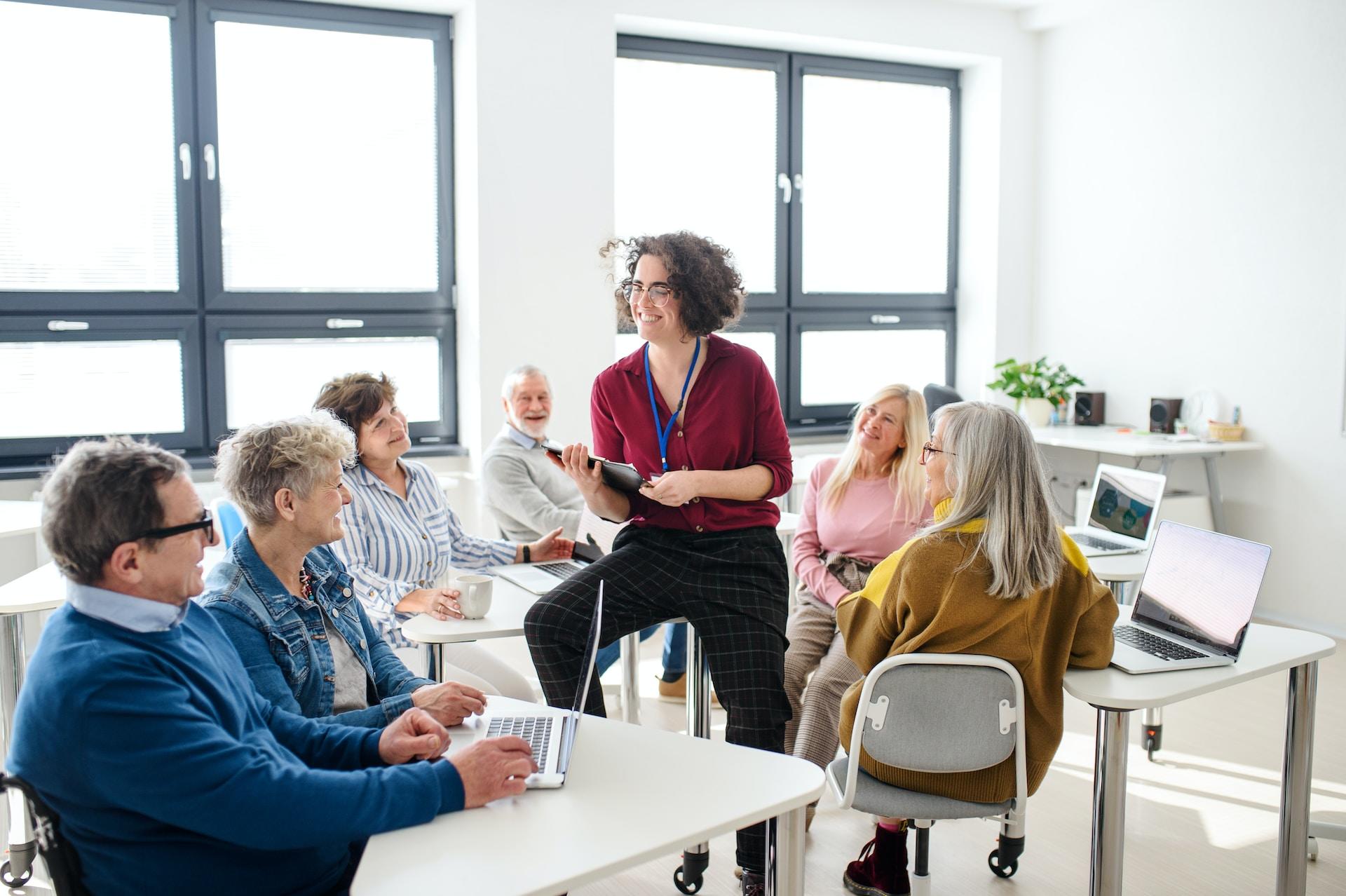 A group of seniors sit at white tables in an informal classroom, each pair with an open laptop before them, as an instructor wearing a dark red top teaches them.