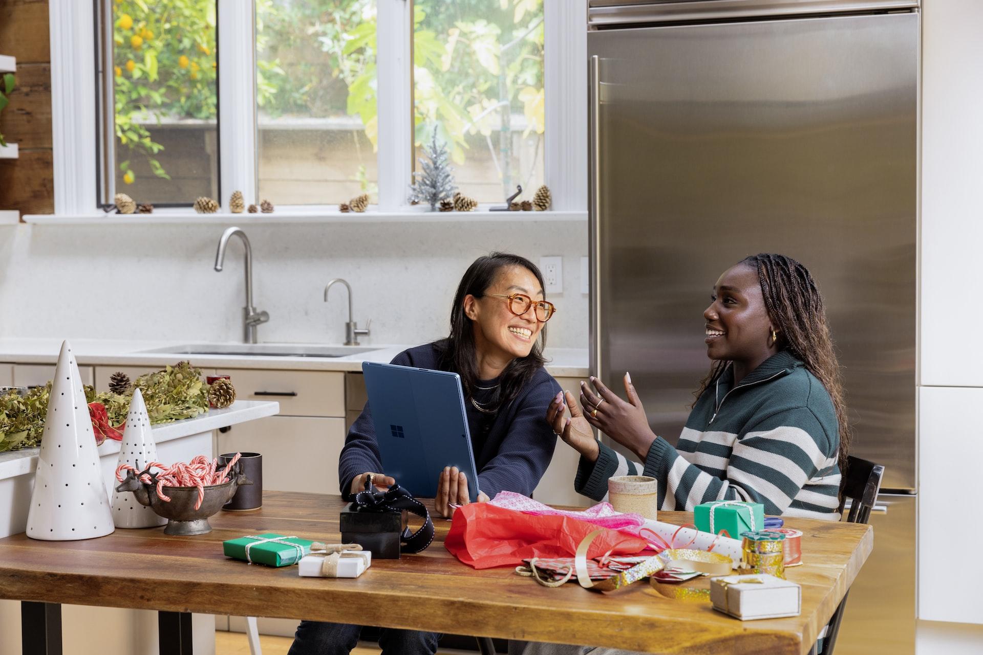 Two women sit at a kitchen table talking animatedly about something they see on a digital tablet.