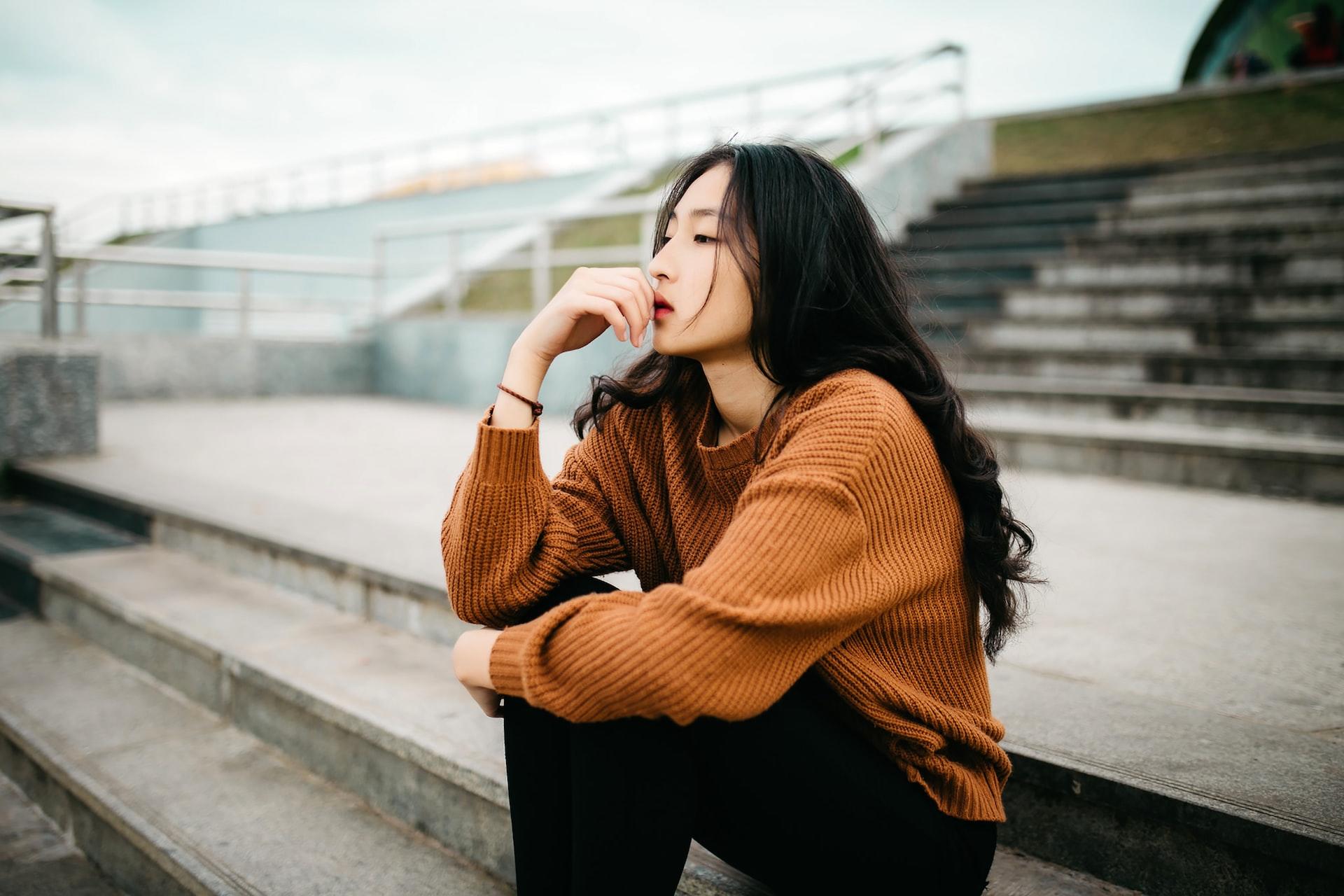 A woman with long hair, wearing a brown top and dark trousers sits reflectively on a concrete stadium riser.