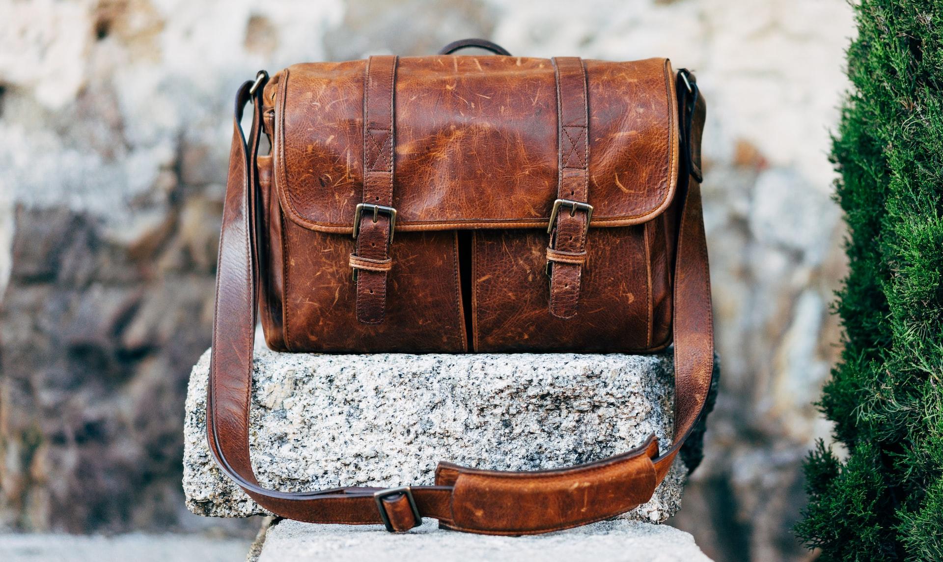 A light brown leather satchel with two buckles and a shoulder strap sits on a grey stone next to an evergreen tree.