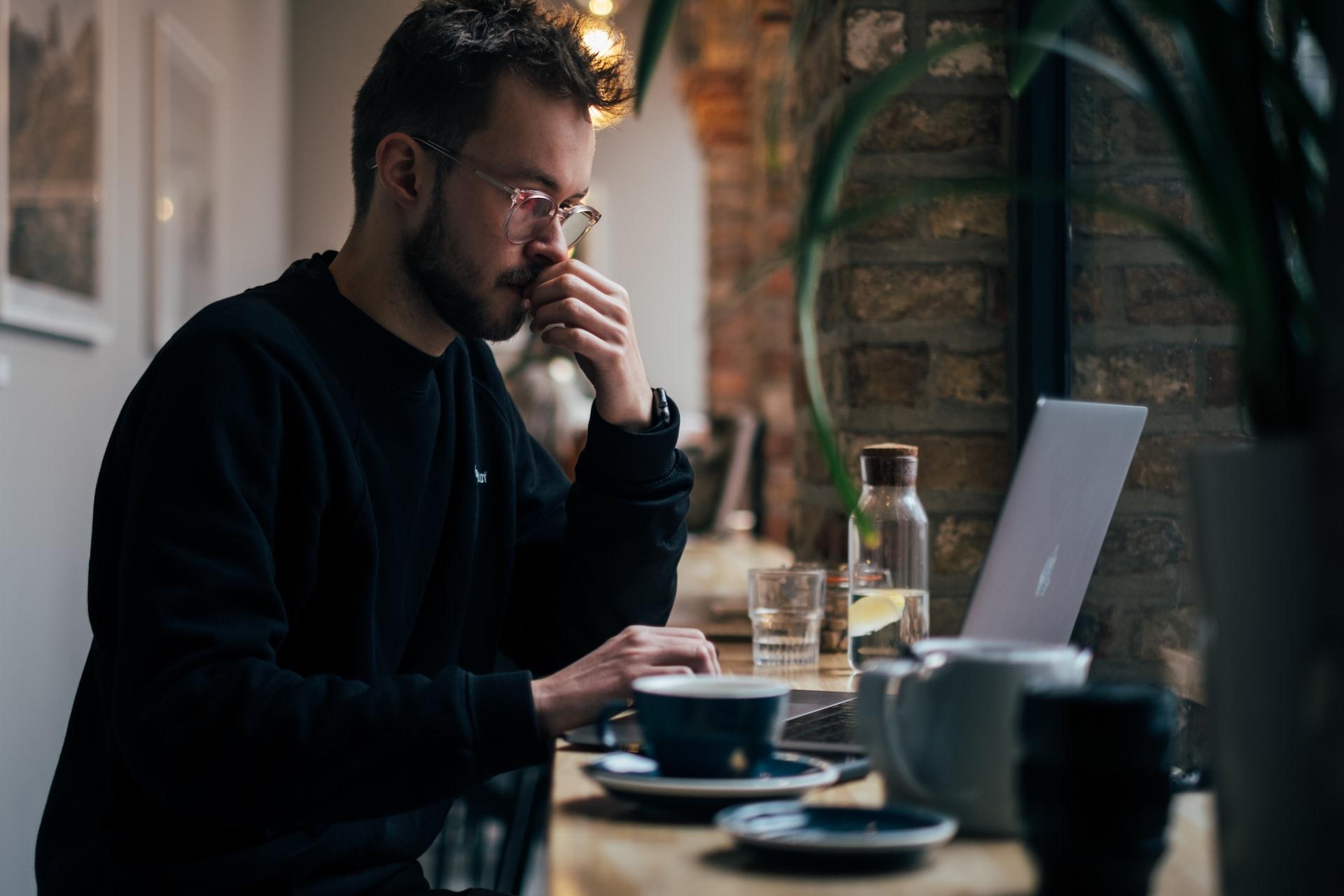 A man in a black jumper looks pensively at his open laptop computer while sitting in a cafe, with a clear glass bottle and a cup within his reach.